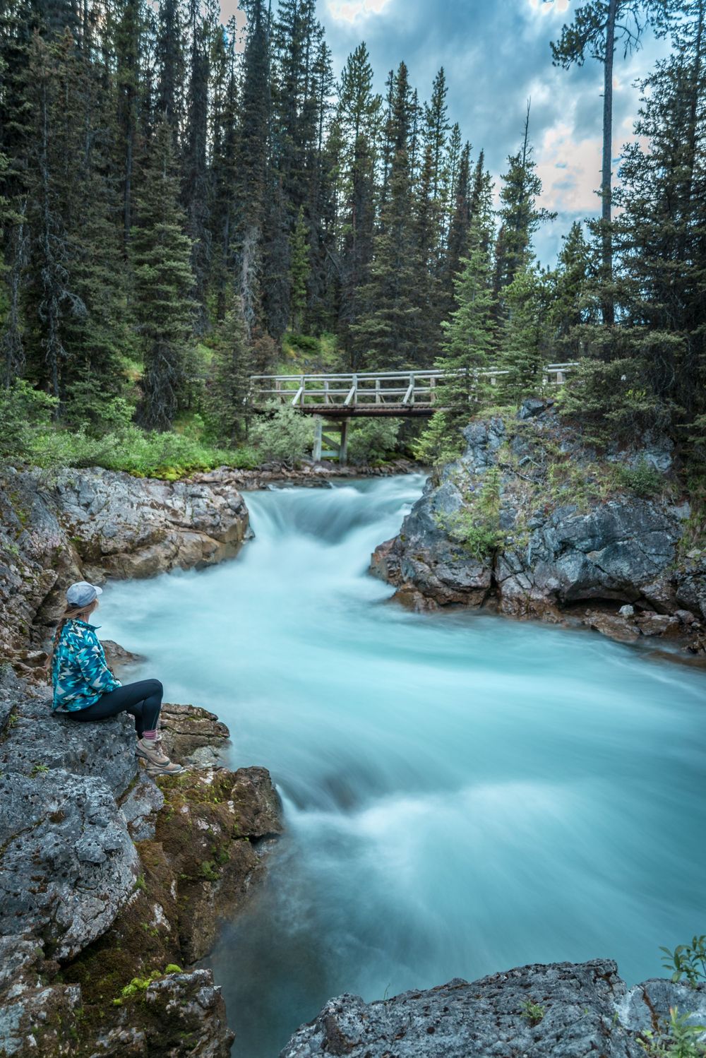 Turquoise glacial river flows beneath wooden footbridge surrounded by evergreen forest in mountain wilderness.