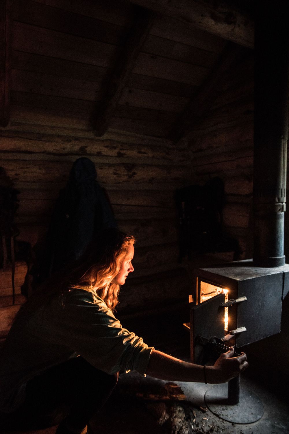 Person tending to a wood stove inside a rustic log cabin illuminated by warm firelight.