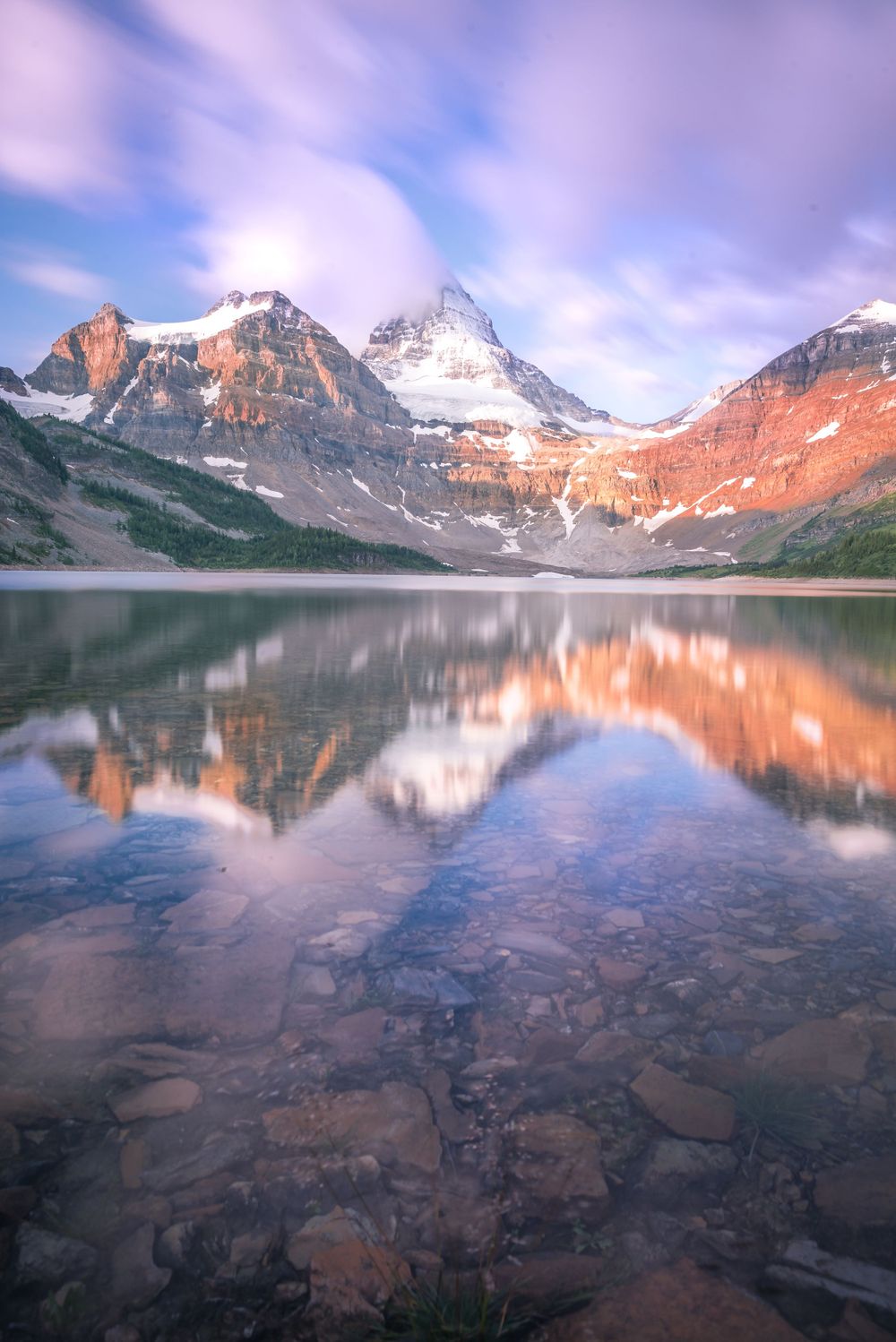 Mountain peaks with snow and reddish cliffs perfectly mirrored in a clear alpine lake at sunrise.