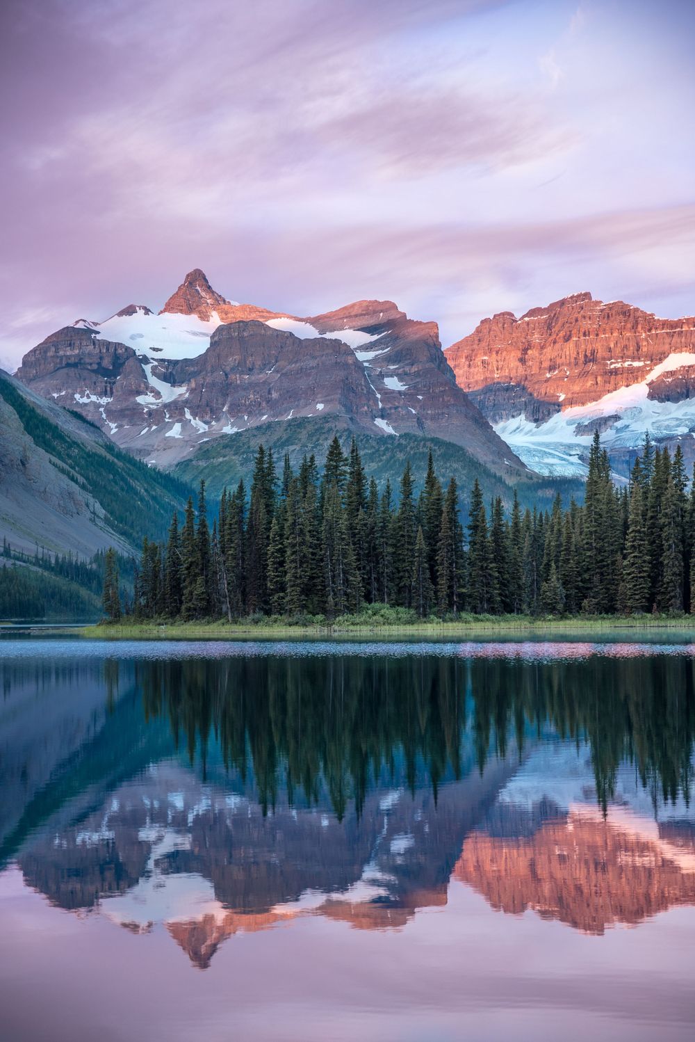 Snow-capped mountain peaks reflect perfectly in calm alpine lake at sunset with evergreen forest in foreground.
