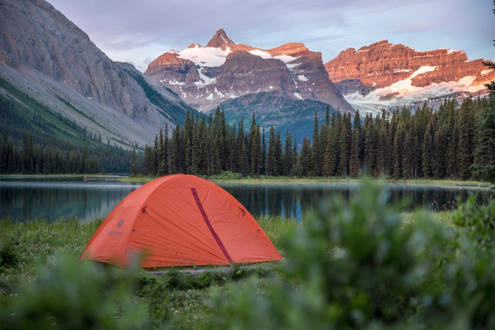 Orange tent glows beside alpine lake at sunset with dramatic mountain peaks in background.