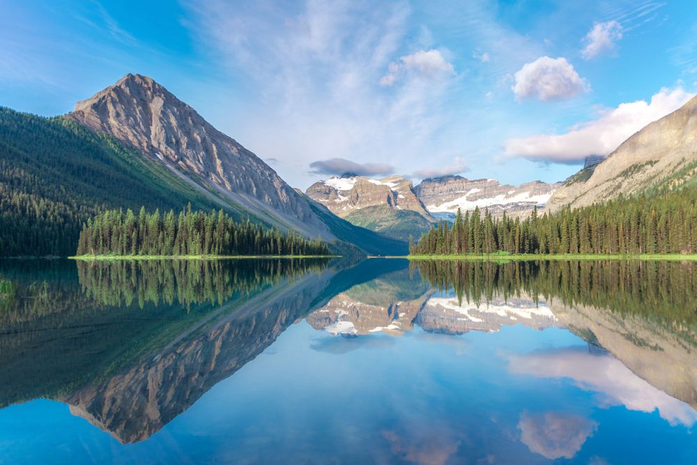 Mirror-like mountain lake reflects symmetrical peaks and evergreen forest under bright blue sky.