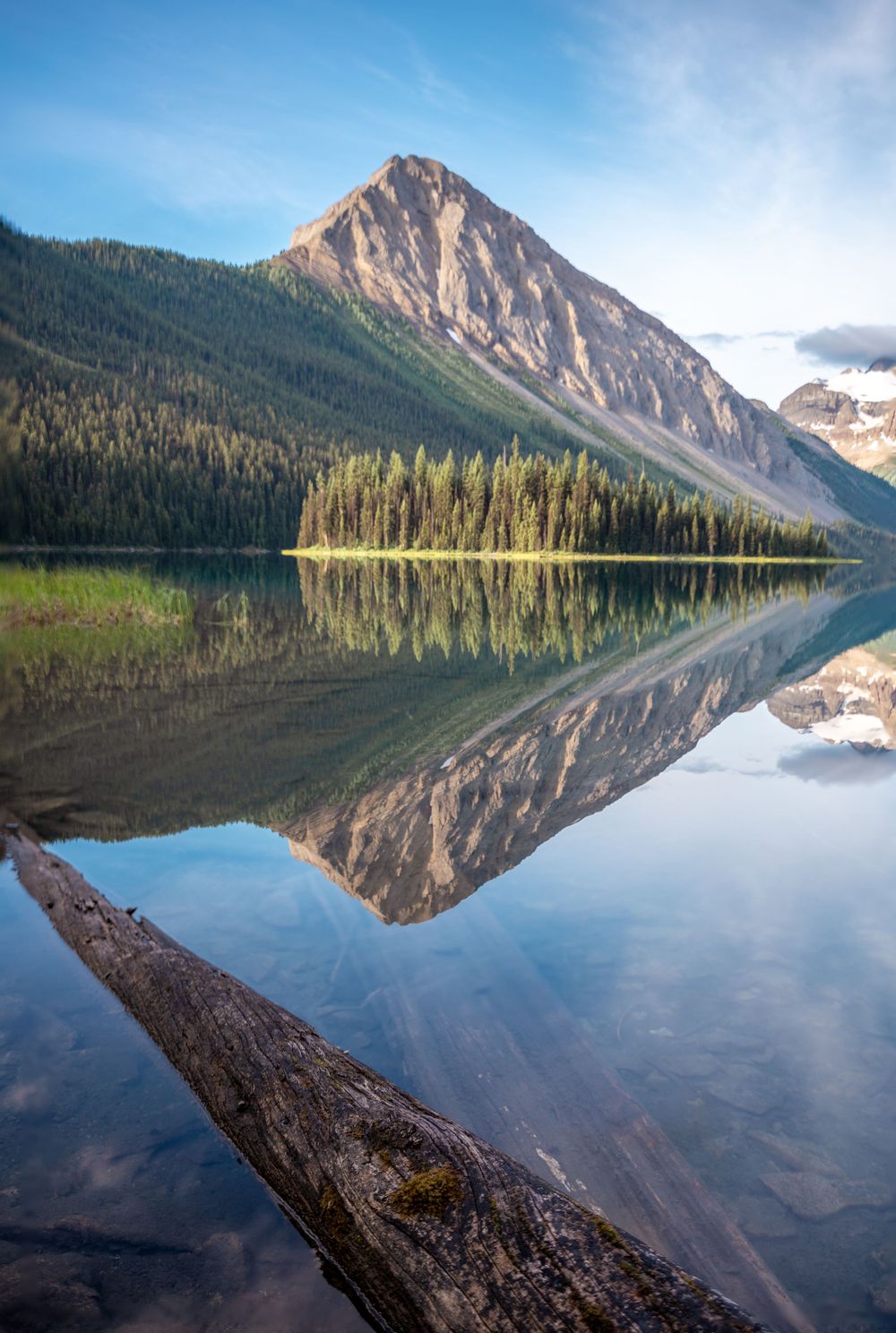 Mountain peak reflects perfectly in calm lake waters surrounded by dense forest on a clear blue day.