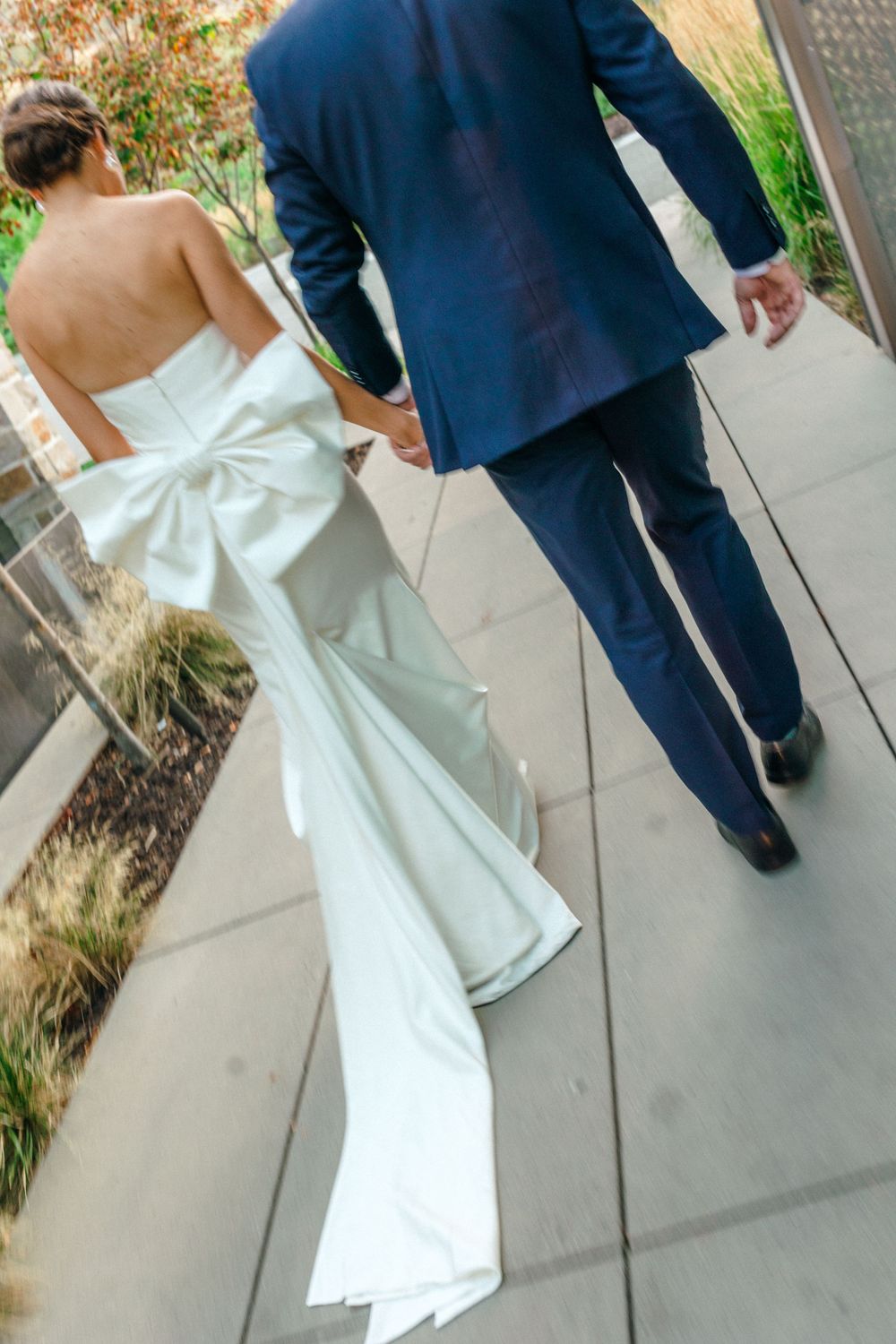 A couple walking shows off the dramatic bow detail on the back of an elegant white wedding gown.