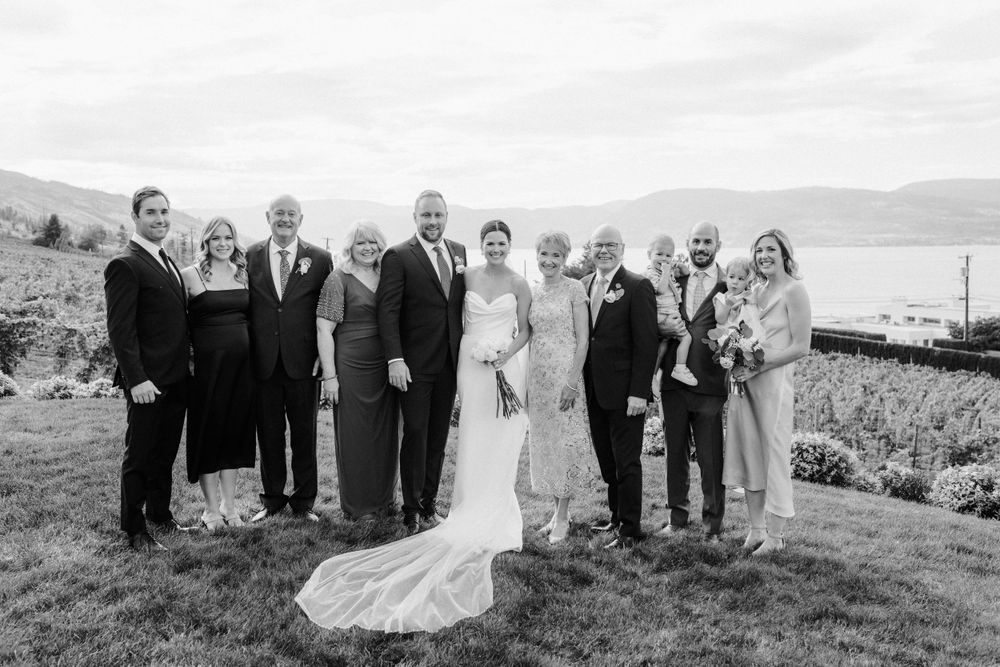 A formal group stands together on a grassy hillside overlooking water on a cloudy day.