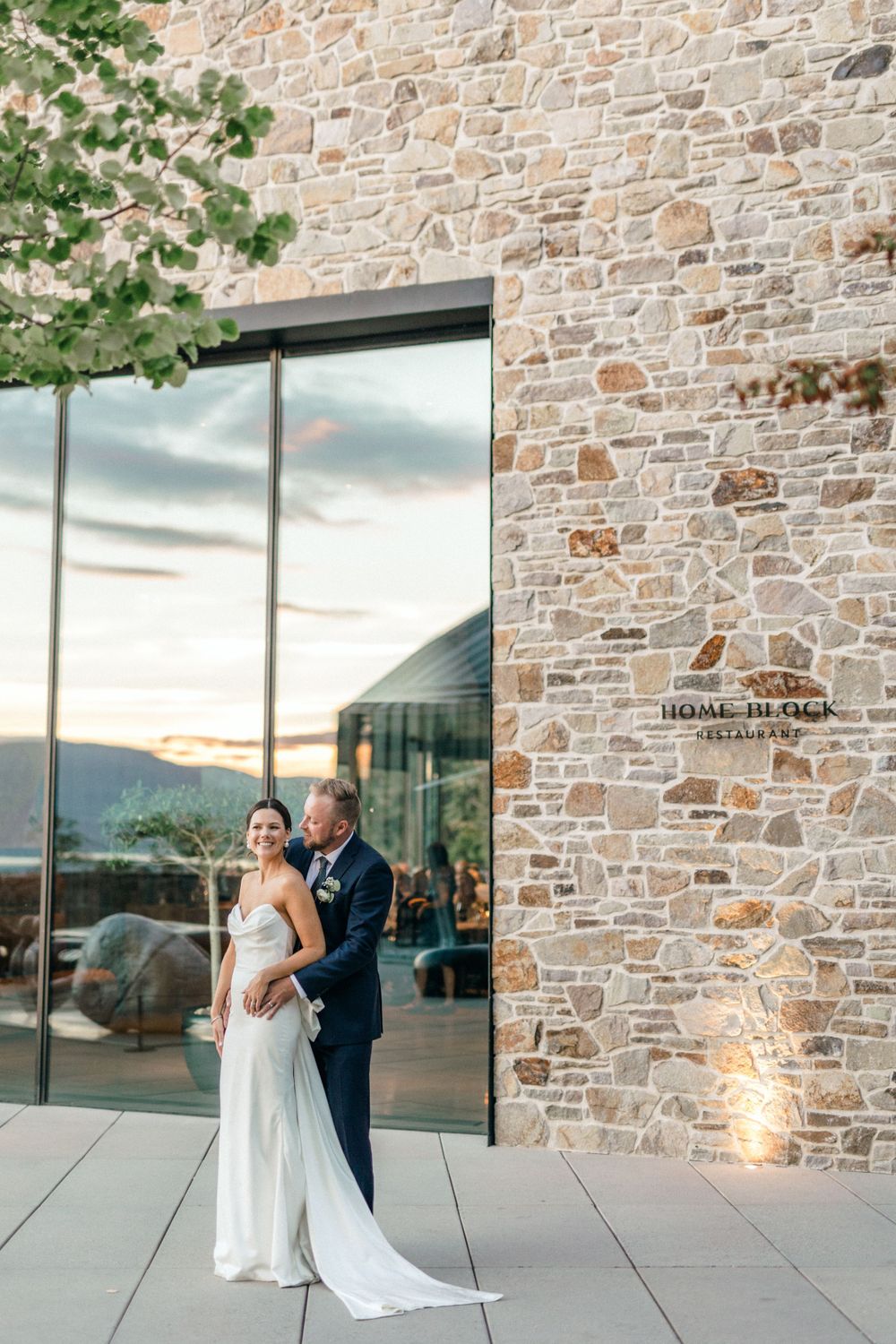 Wedding couple poses against modern stone building with reflective windows at sunset.