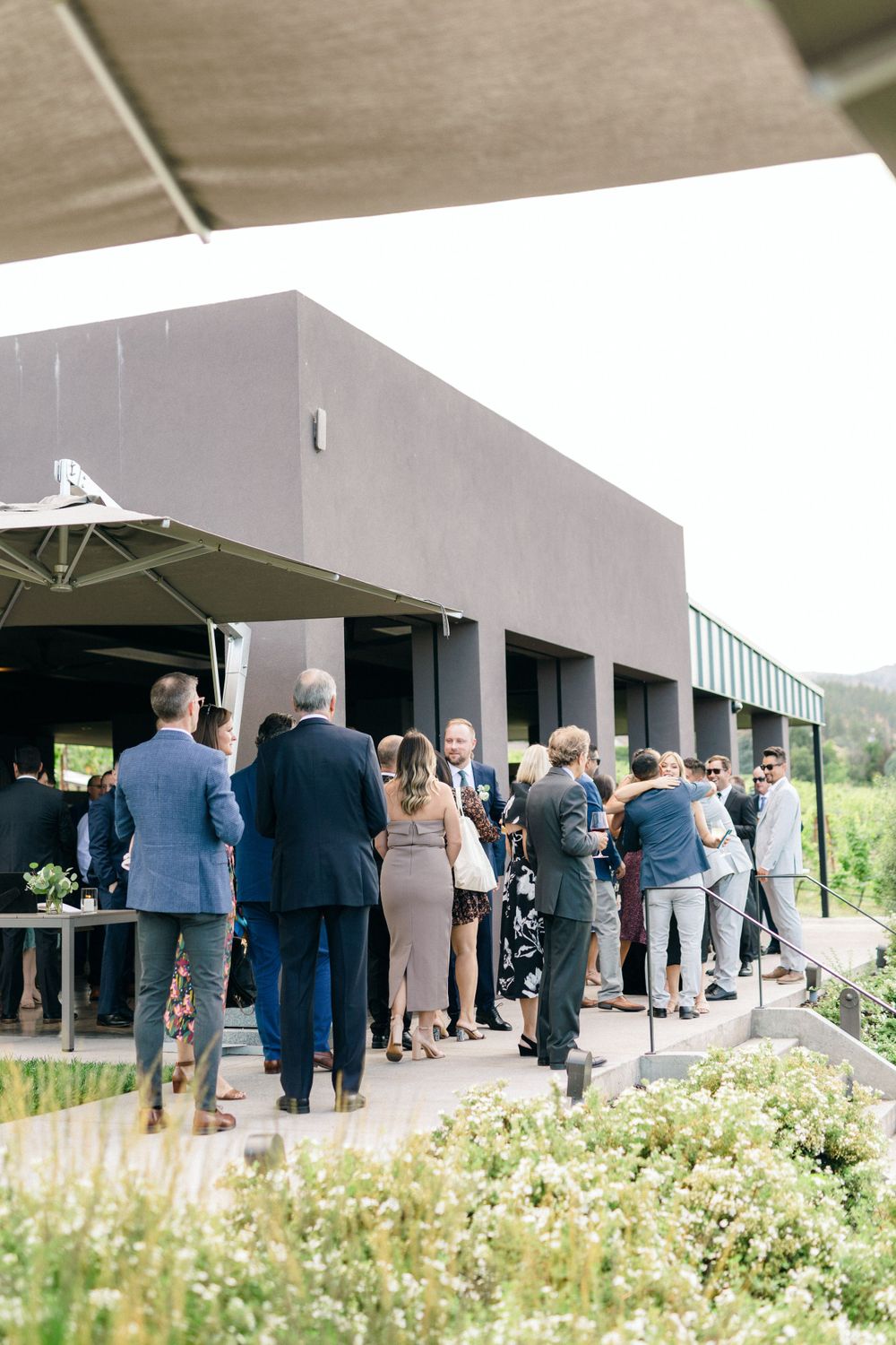 A group of well-dressed guests gathered outside a modern venue building with wildflowers in the foreground.