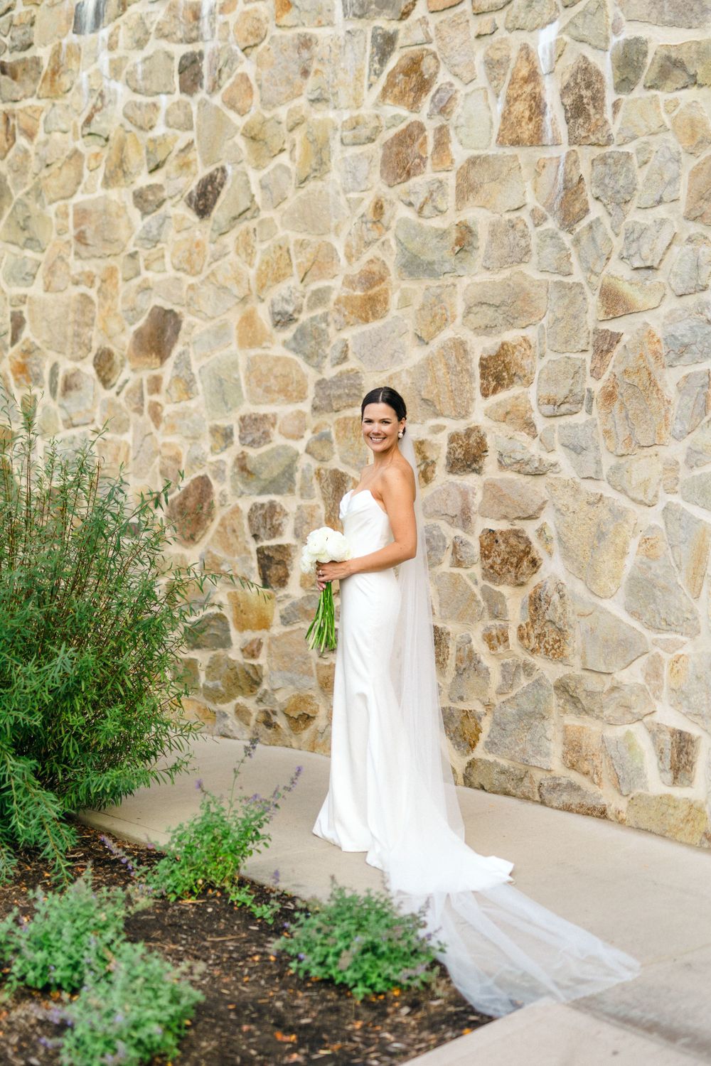 Beautiful bride in flowing white dress poses against rustic stone wall with garden foliage at cedar creek winery in the Okanagan.