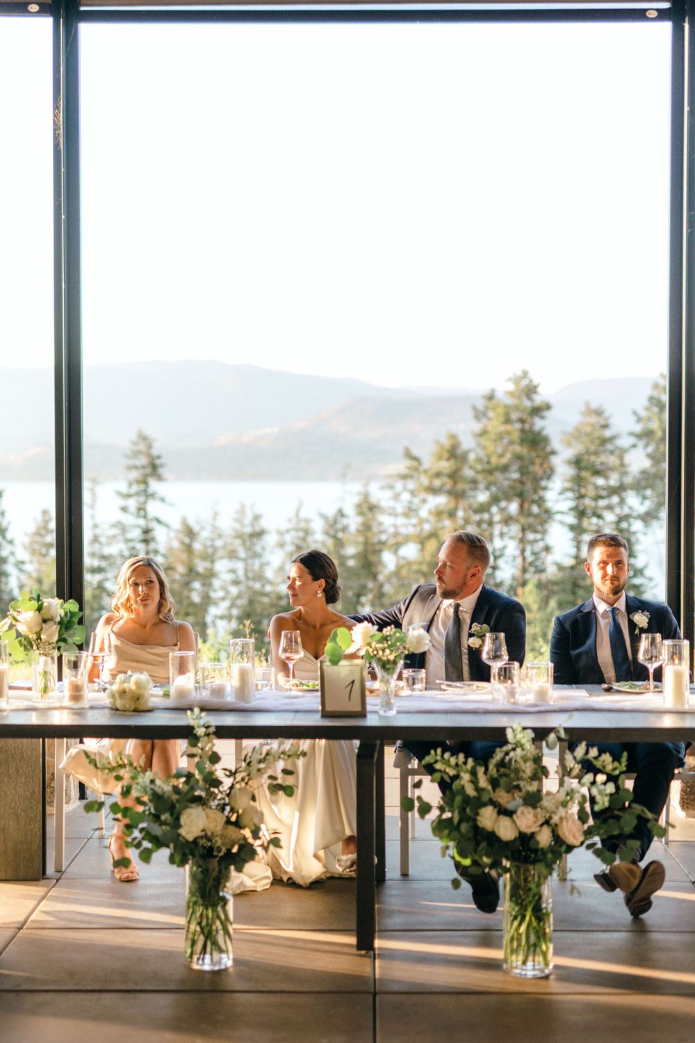 Wedding party seated at head table with mountain lake view and elegant white floral centerpieces at sunset.