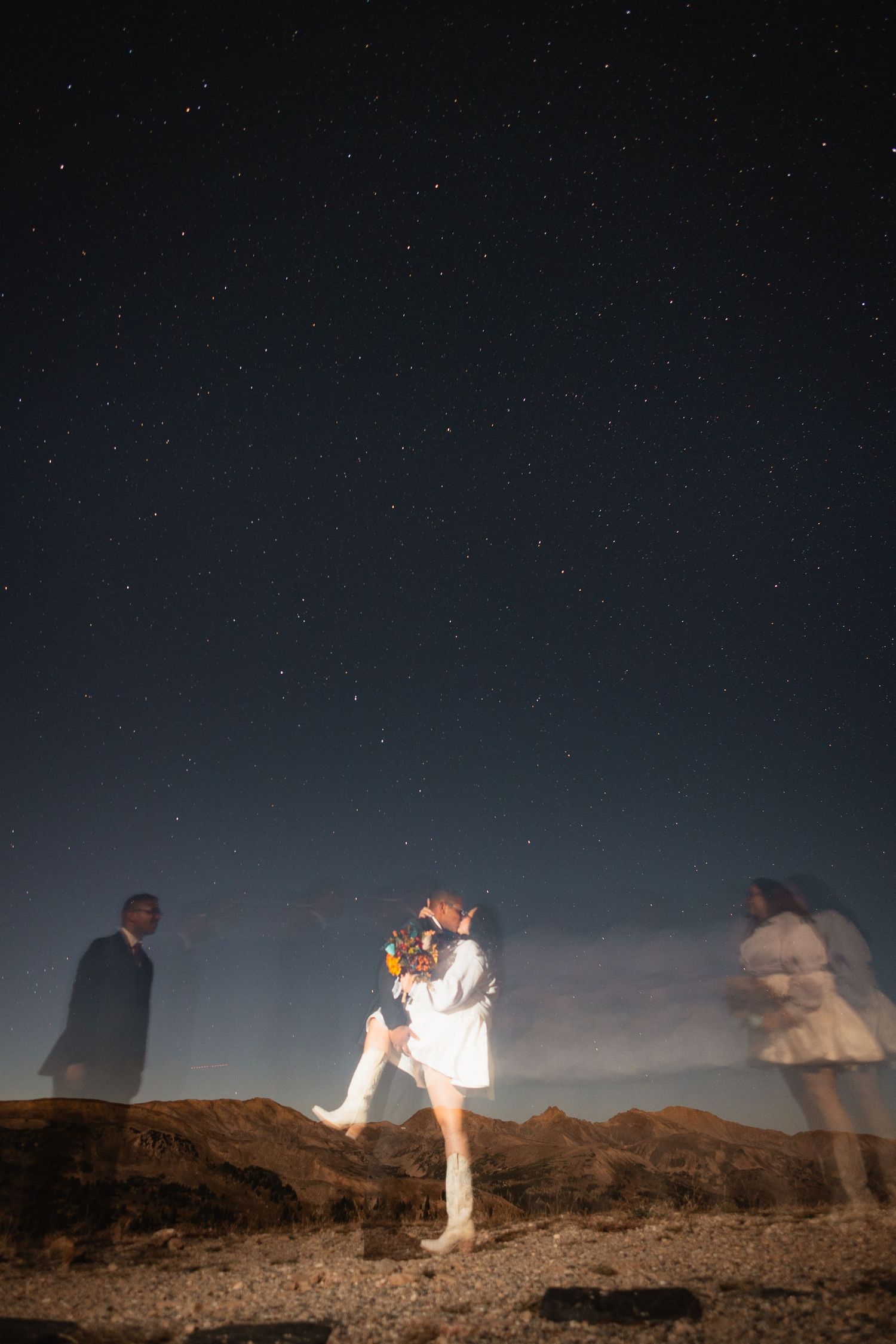 A long exposure night shot captures blurred figures on a desert hill under a starry sky with ghostly motion trails.
