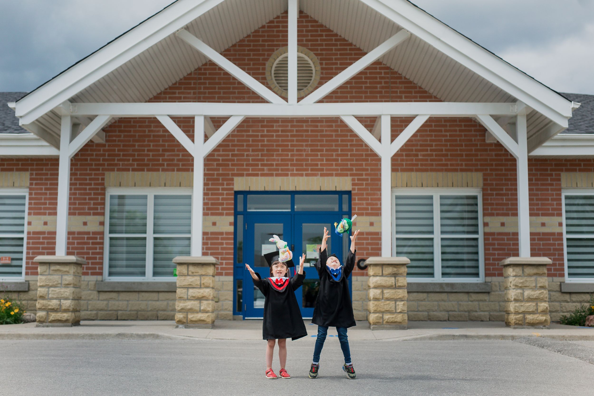 Graduates celebrate in front of a brick building with white trim and a blue door on a cloudy day.