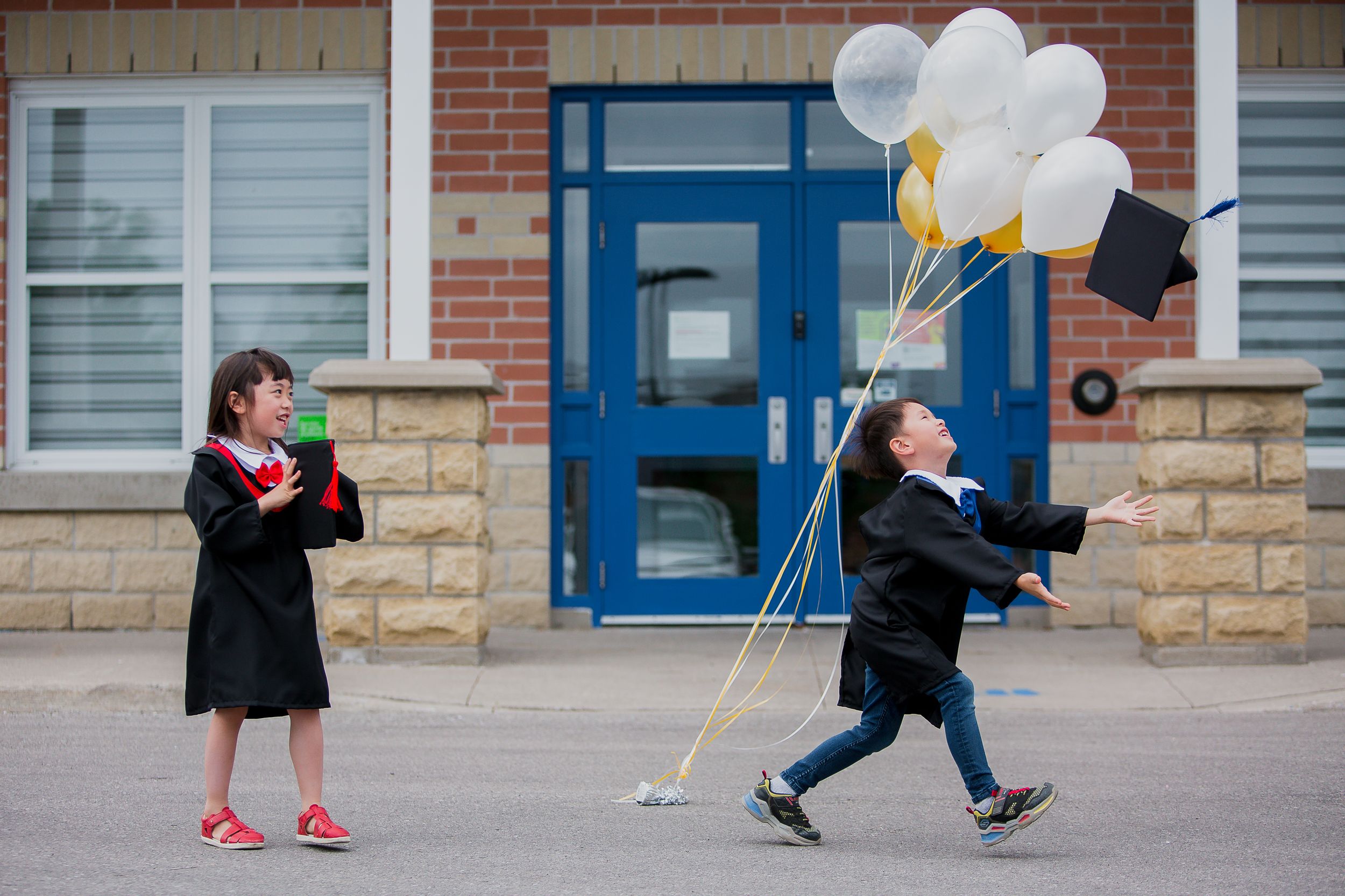 Two graduates celebrate outside a building with white balloons and graduation attire during a graduation ceremony.