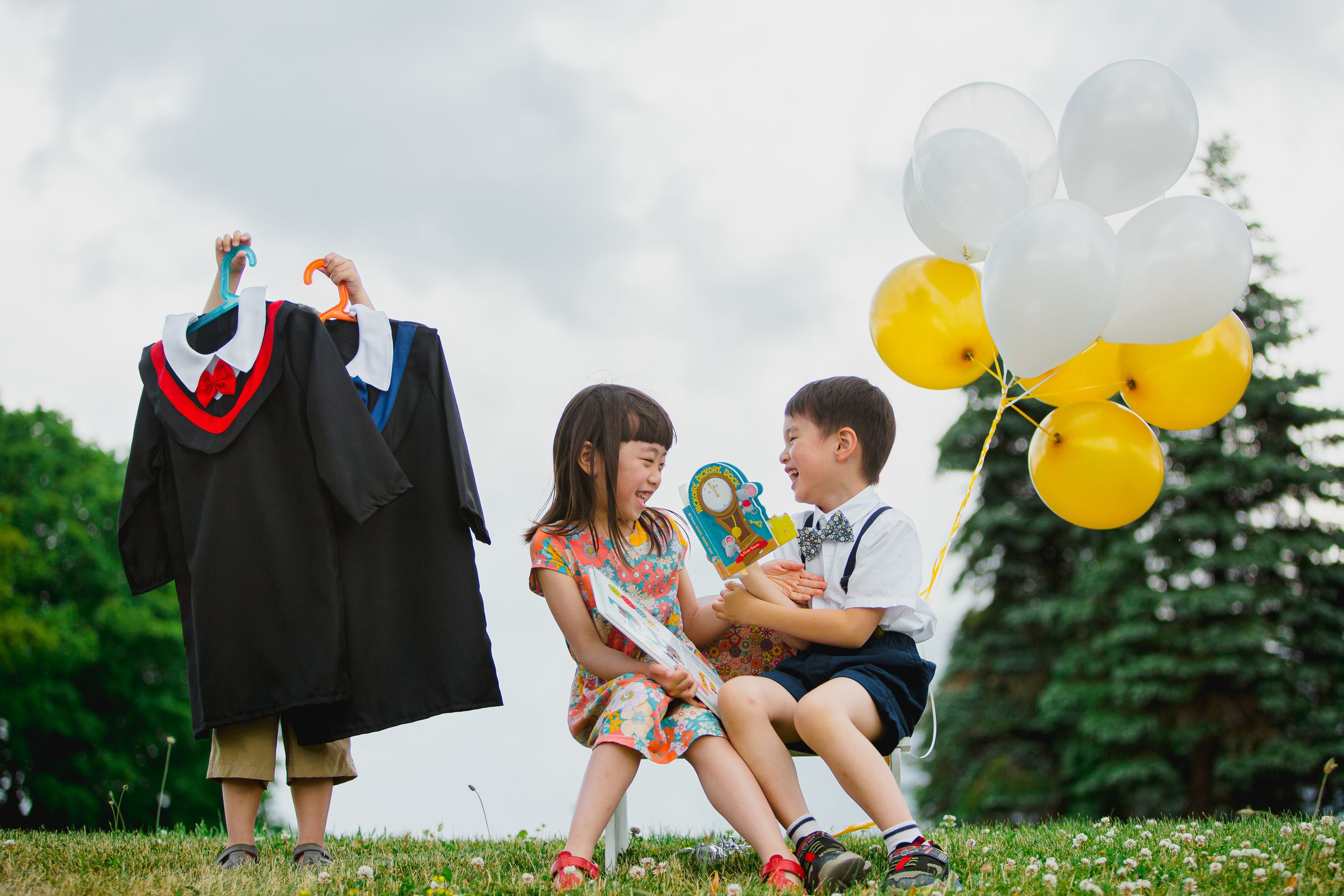 Academic graduation celebration outdoors with diploma gown, children and yellow balloons against cloudy sky.