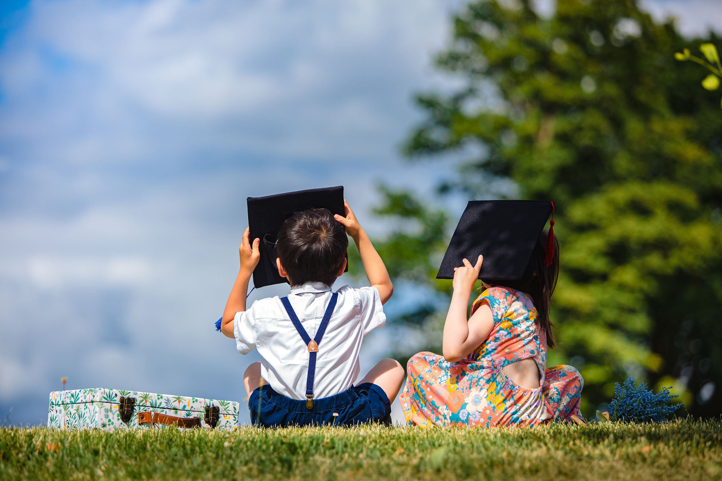 Two students in graduation caps sit on grassy hill on sunny day looking at blue sky with trees in background.