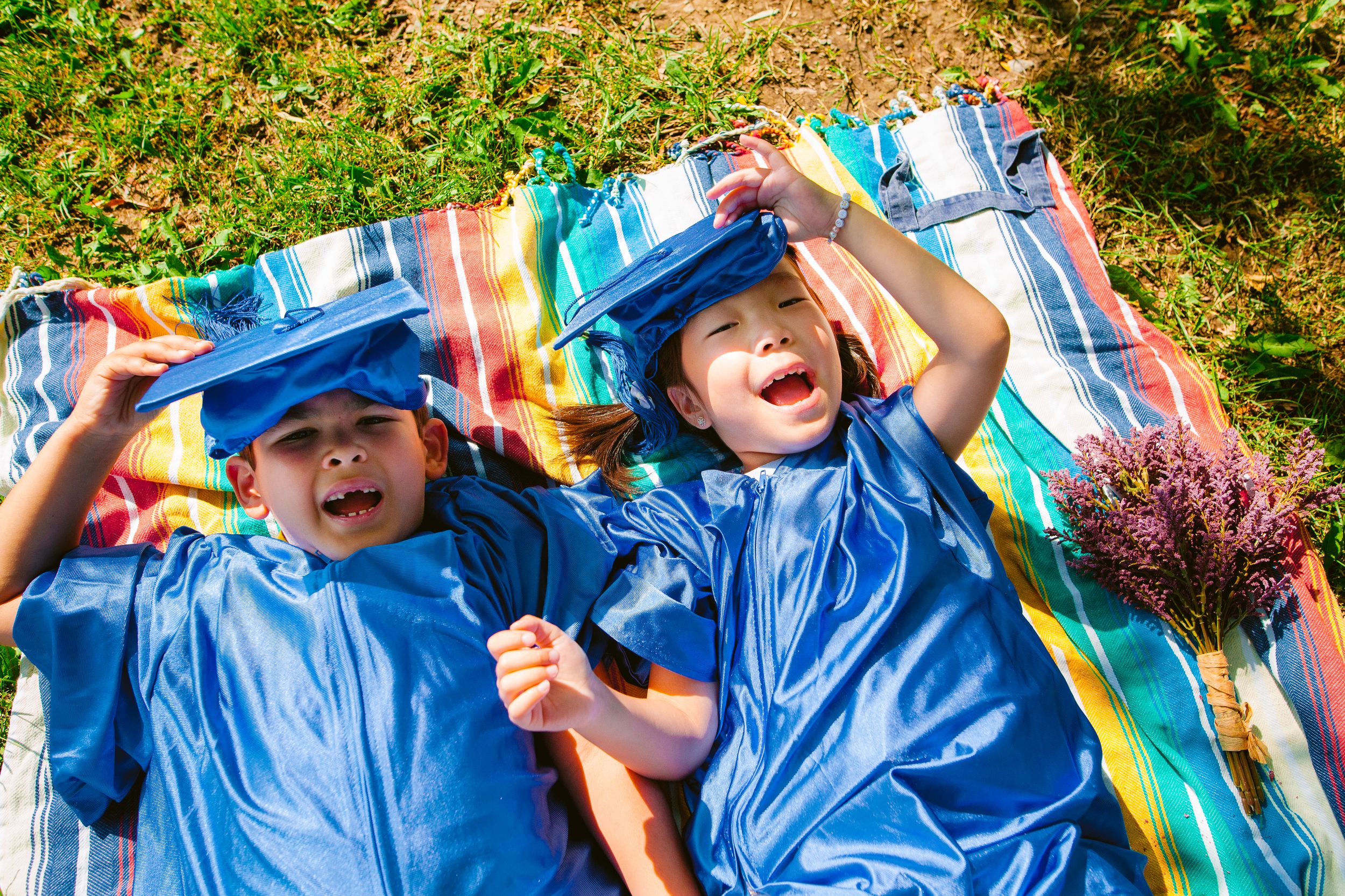 Two children wearing bright blue rain coats lay on a colorful blanket outdoors while smiling and playing together.