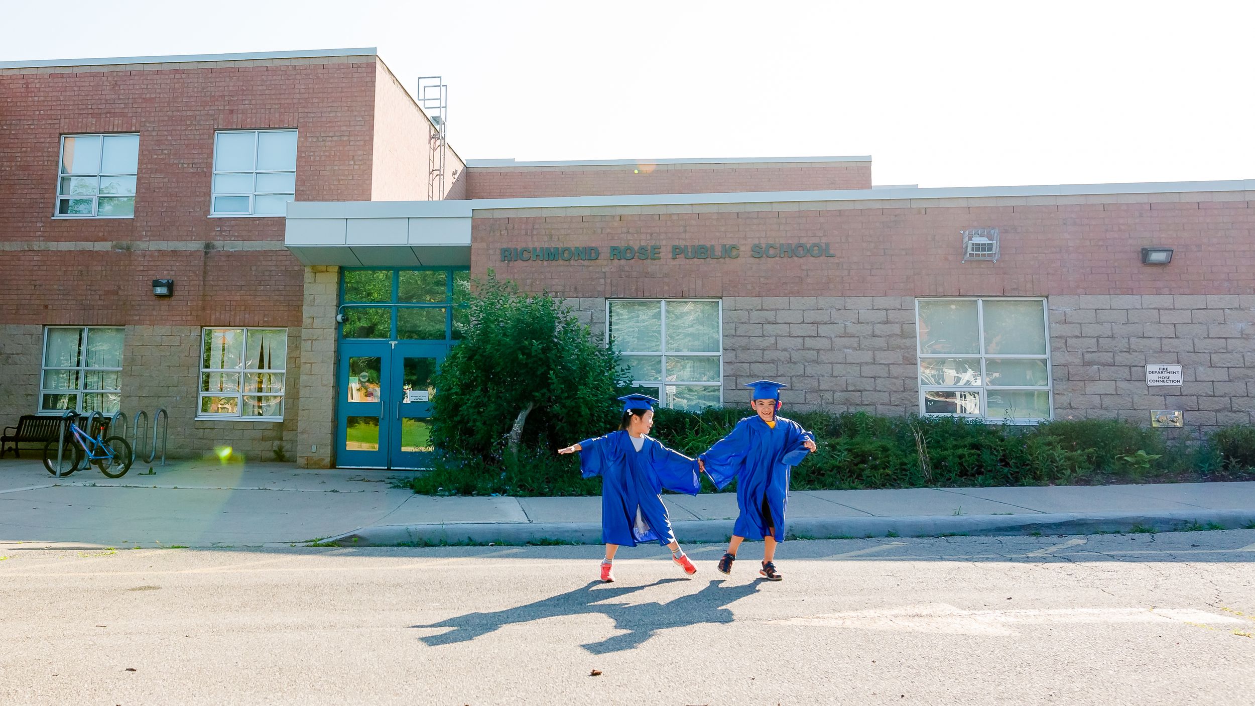 Two people in blue outfits hold hands and cast shadows outside a brick school building entrance.