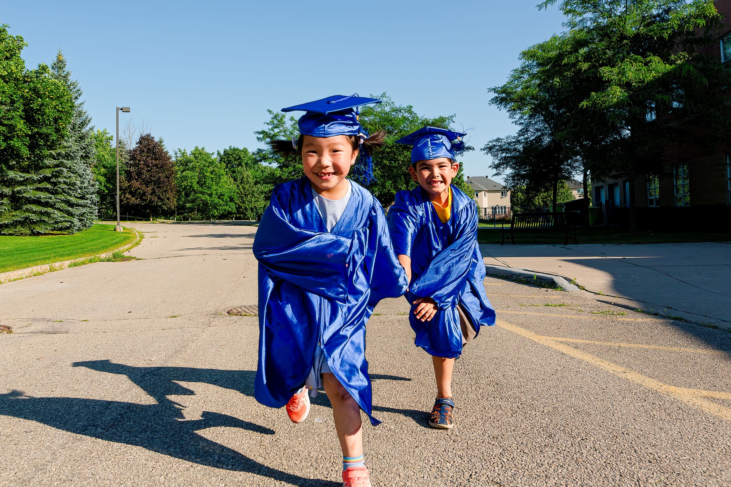 Two young graduates in blue caps and gowns run joyfully down a sunny sidewalk on graduation day.