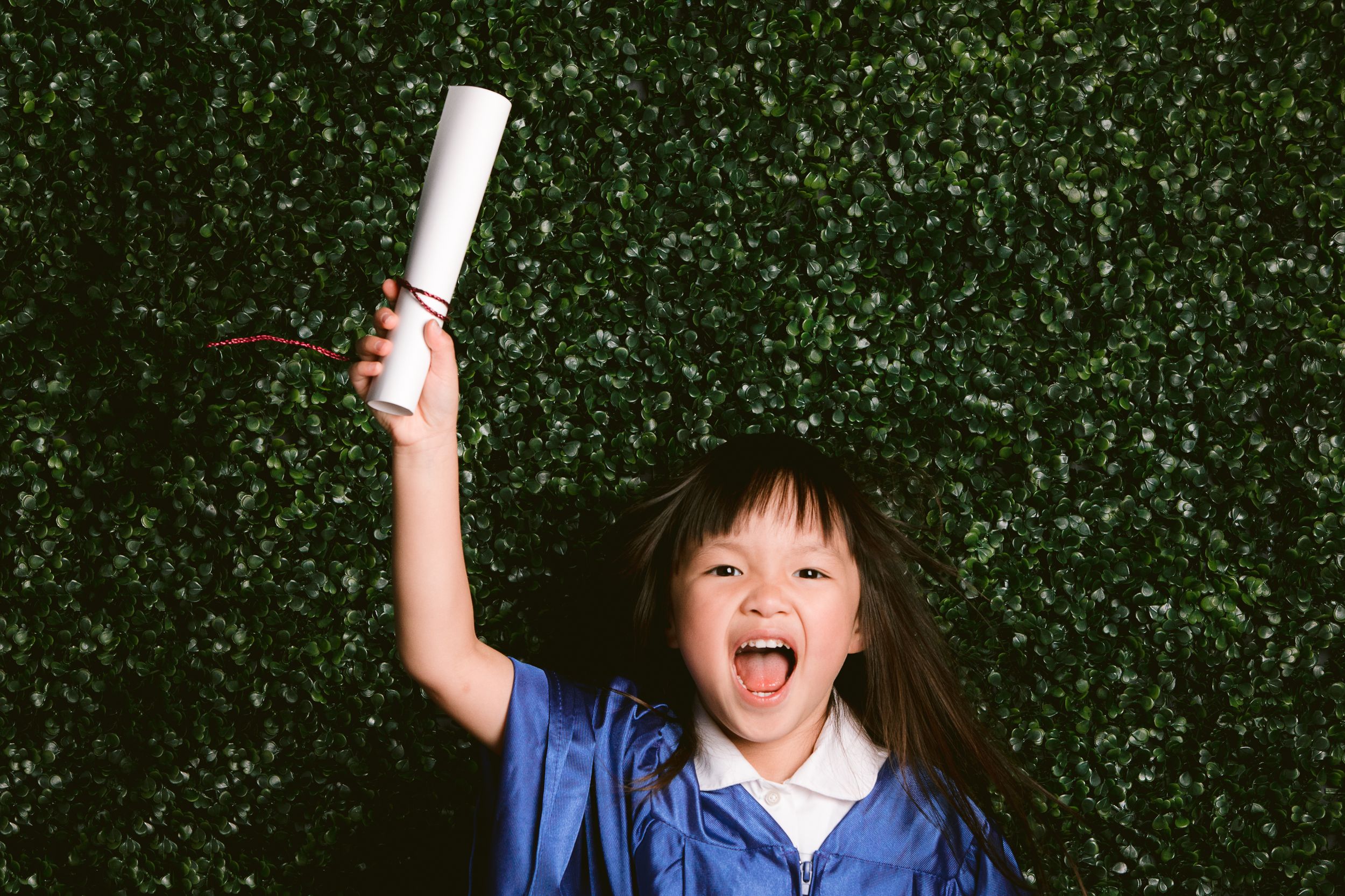 A graduate holds up their diploma while cheerfully celebrating against a green hedge background.
