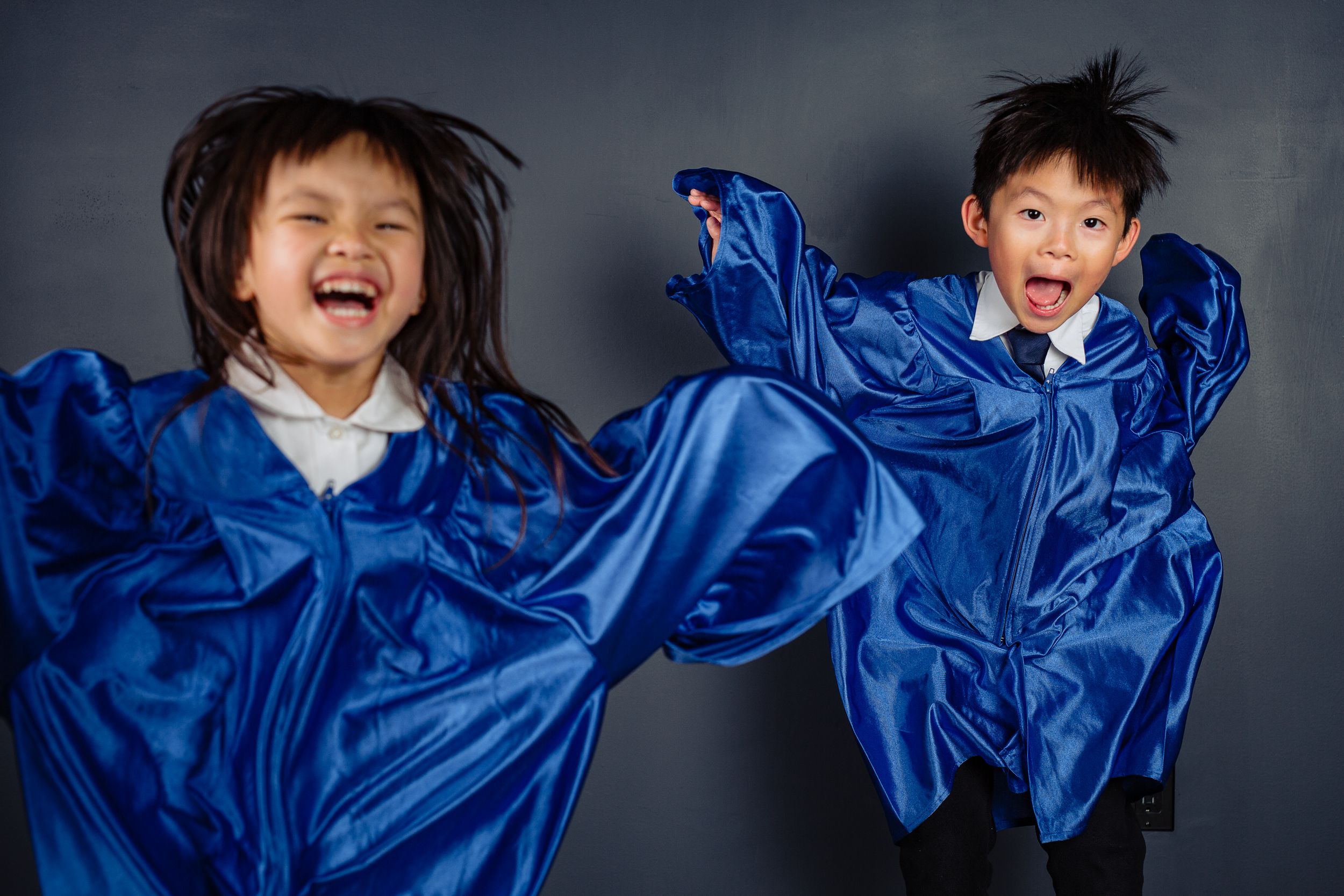 Two energetic children jump and pose in flowing bright blue silk graduation gowns against a gray background.