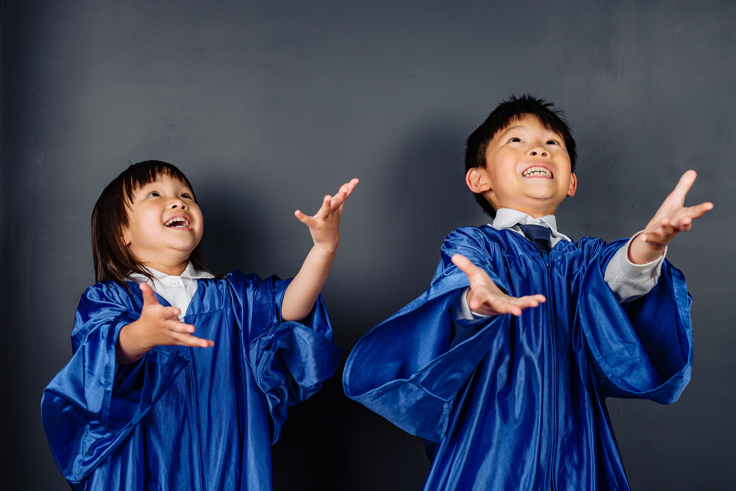 Two graduates in bright blue robes celebrate joyfully with raised arms against a dark background.