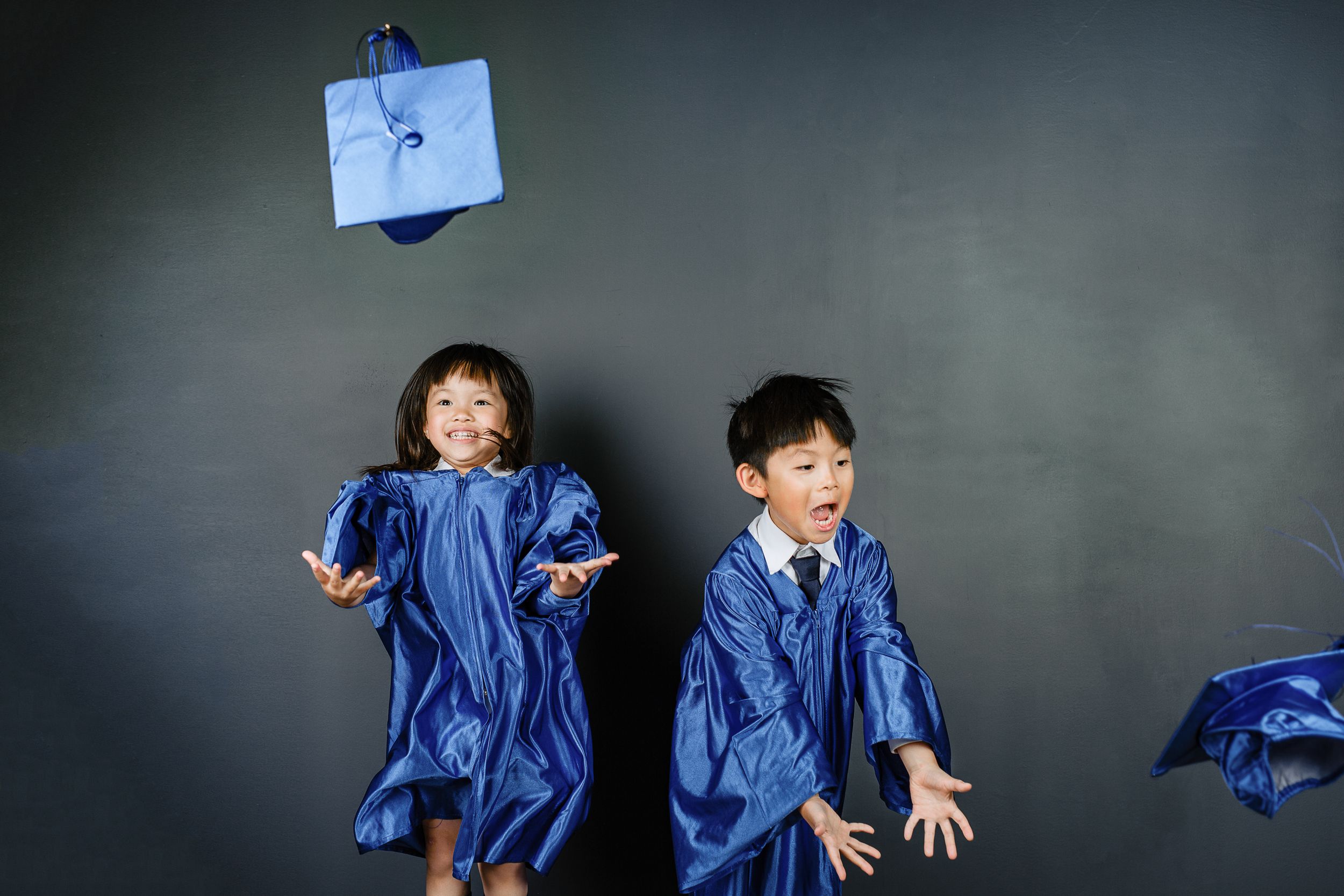 Two children in blue graduation gowns jump playfully against a dark gray background with a floating cap.
