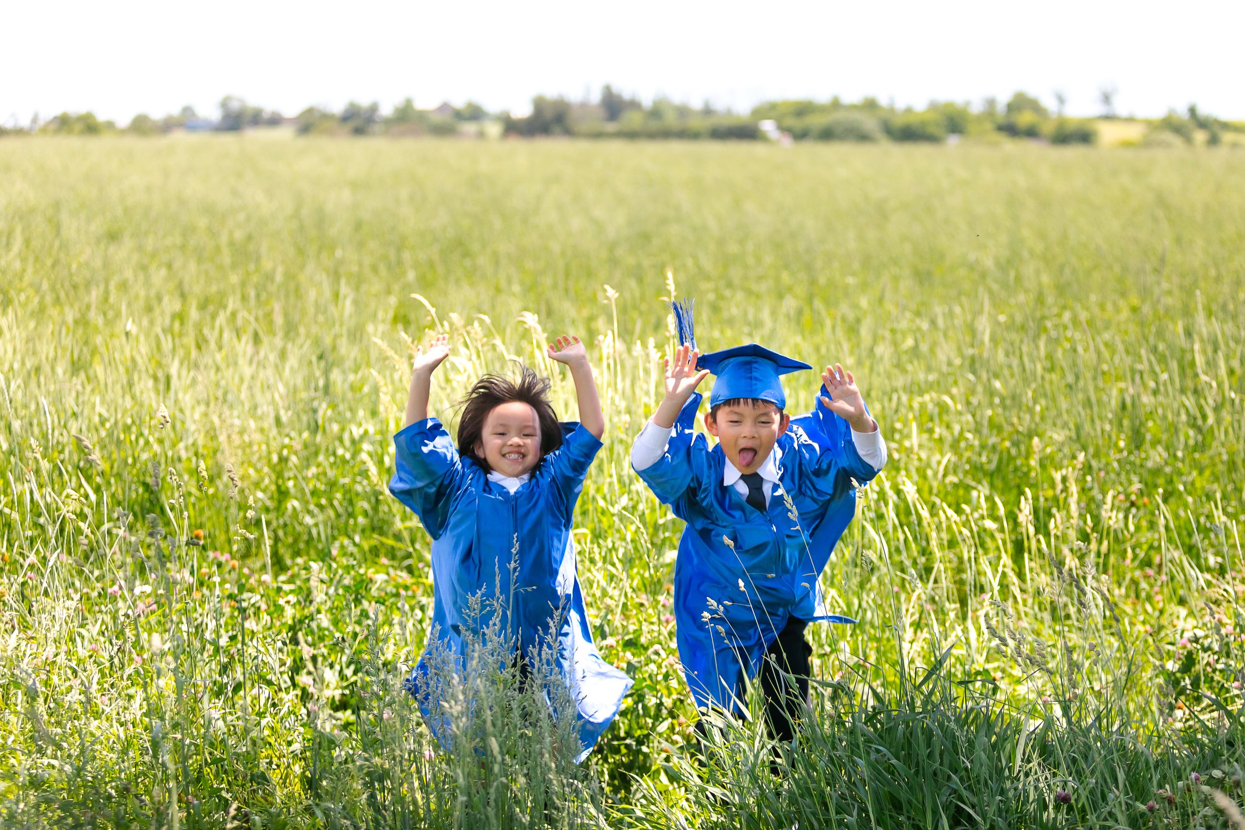 Two young children in blue jackets joyfully playing in a tall grass meadow on a sunny day.