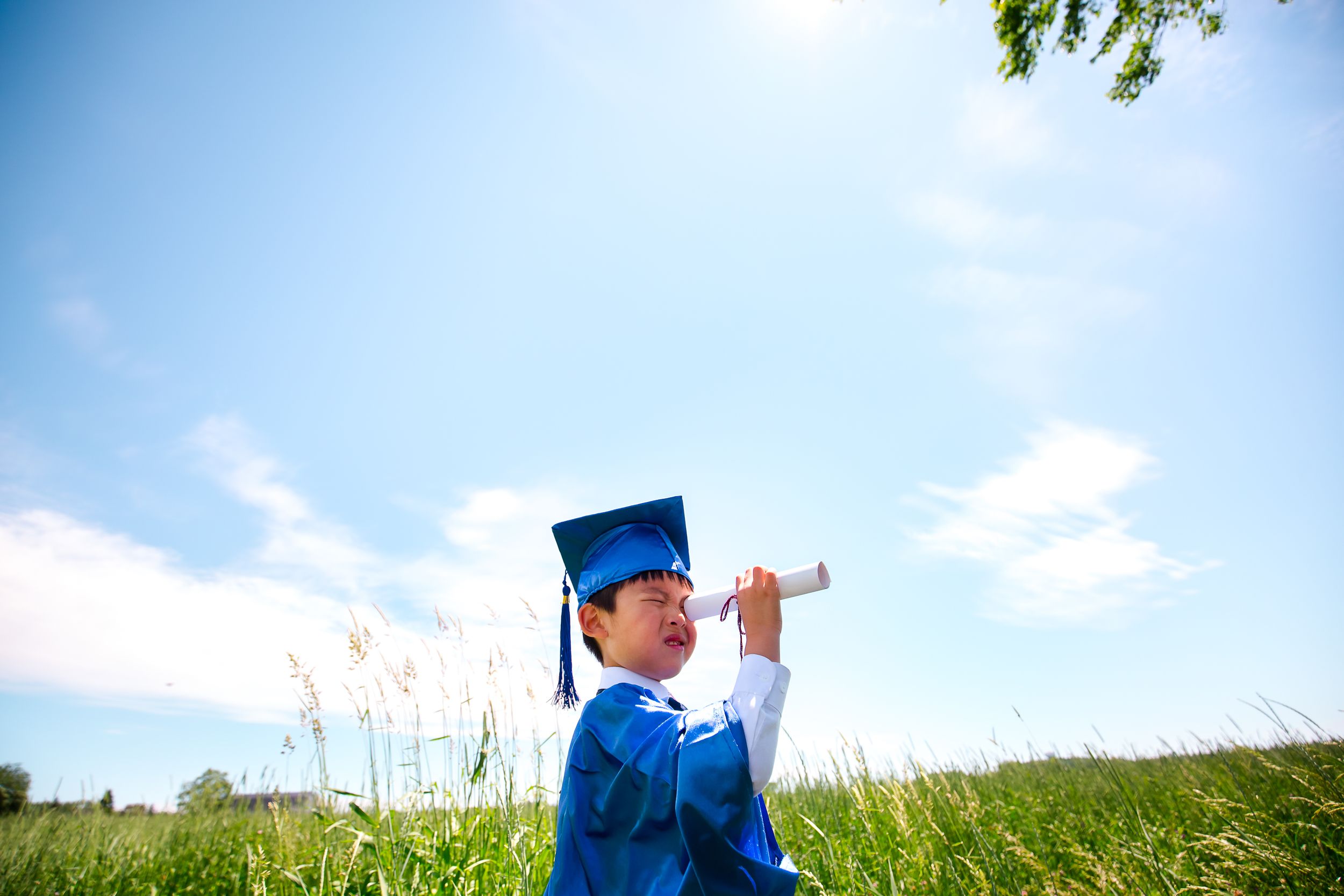 Young graduate in blue cap and gown looks through diploma toward bright blue sky while sitting in grassy field.