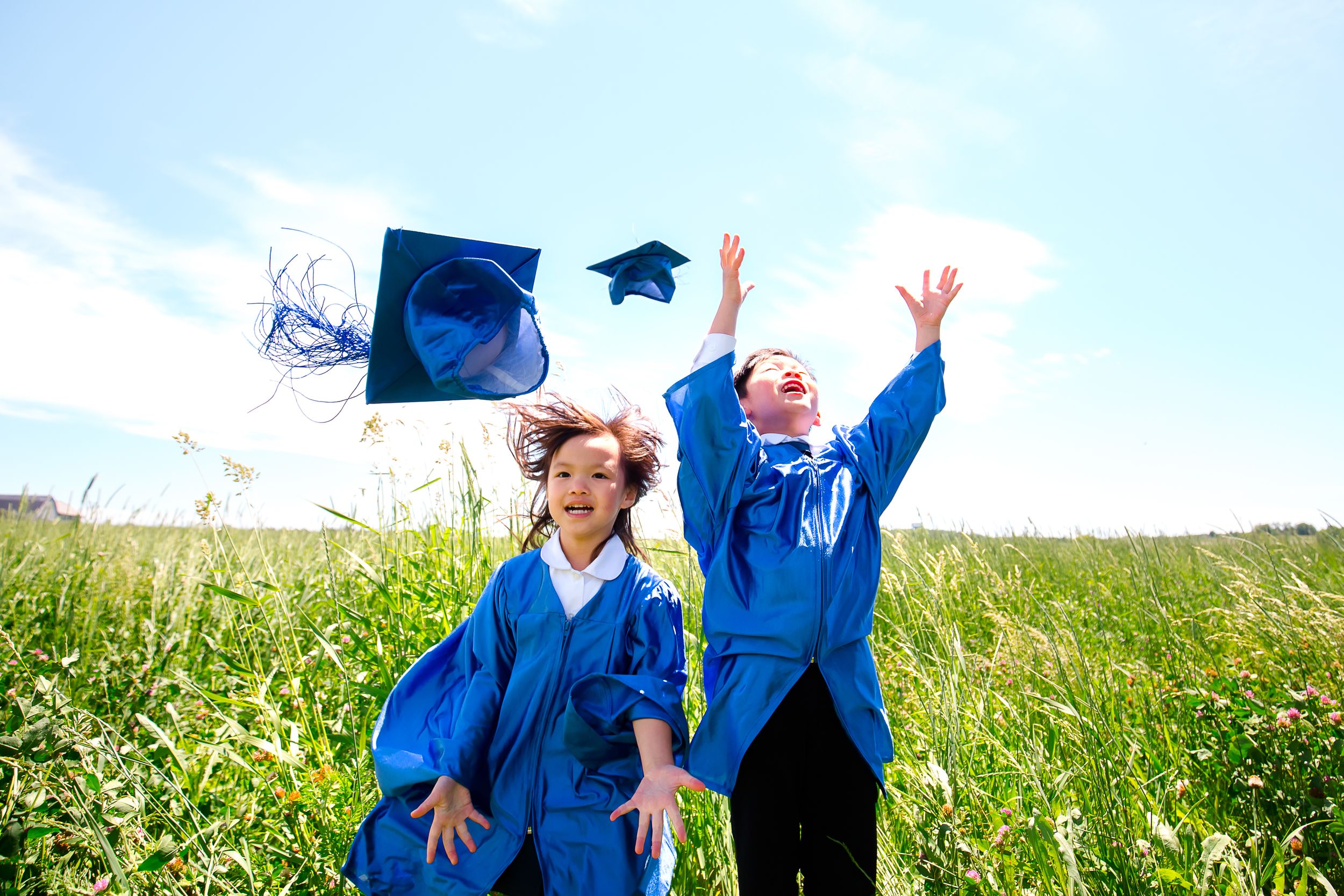 Graduates in bright blue robes celebrate by jumping and tossing caps in a sunny meadow against blue sky.