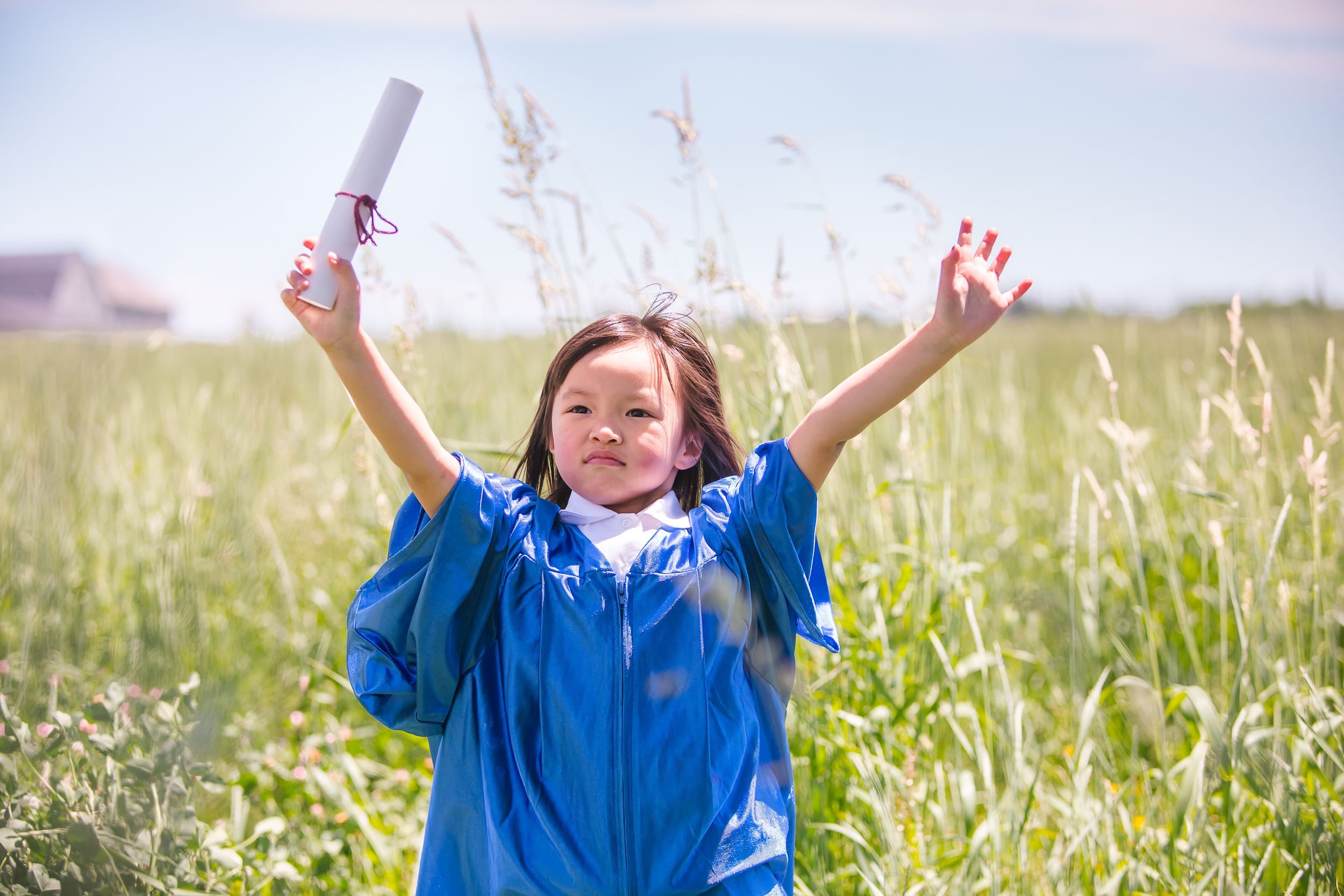 A child in a blue graduation gown celebrates in a sunny meadow with raised arms holding a diploma.