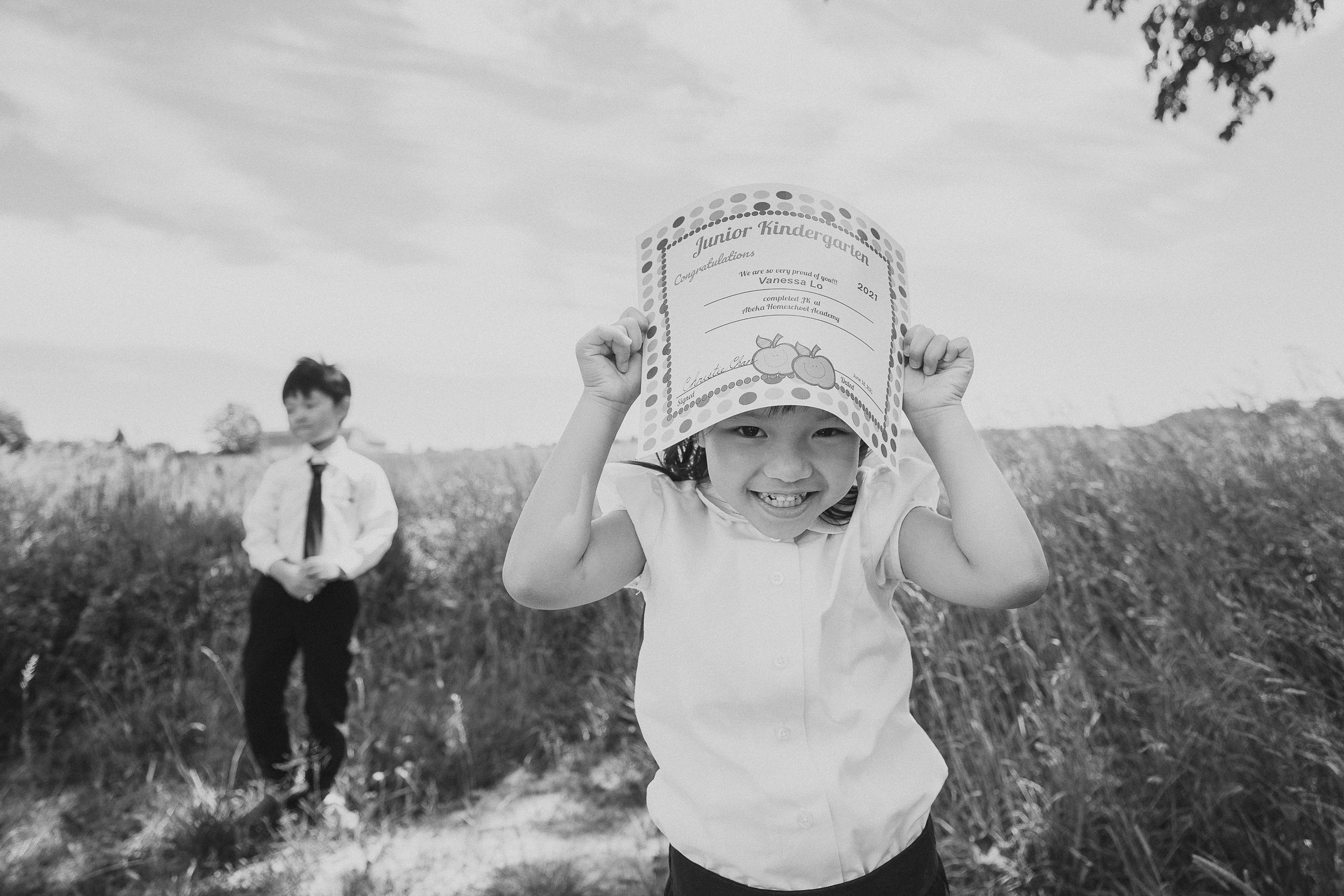 A vintage black and white photograph shows a child in a white shirt playfully holding up a basket in a grassy field.