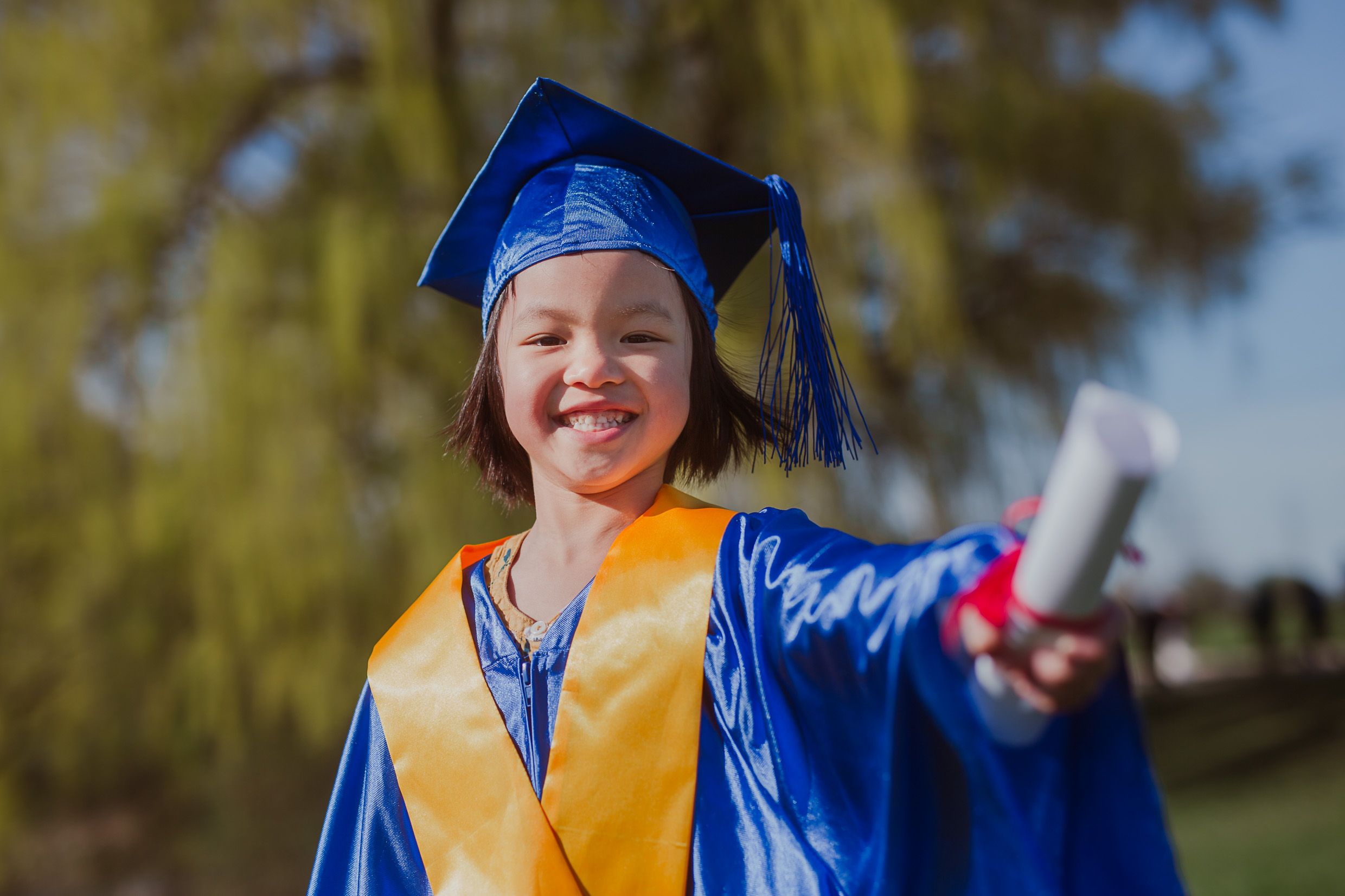 Graduate in blue cap and gown with yellow sash celebrating with diploma at outdoor ceremony.