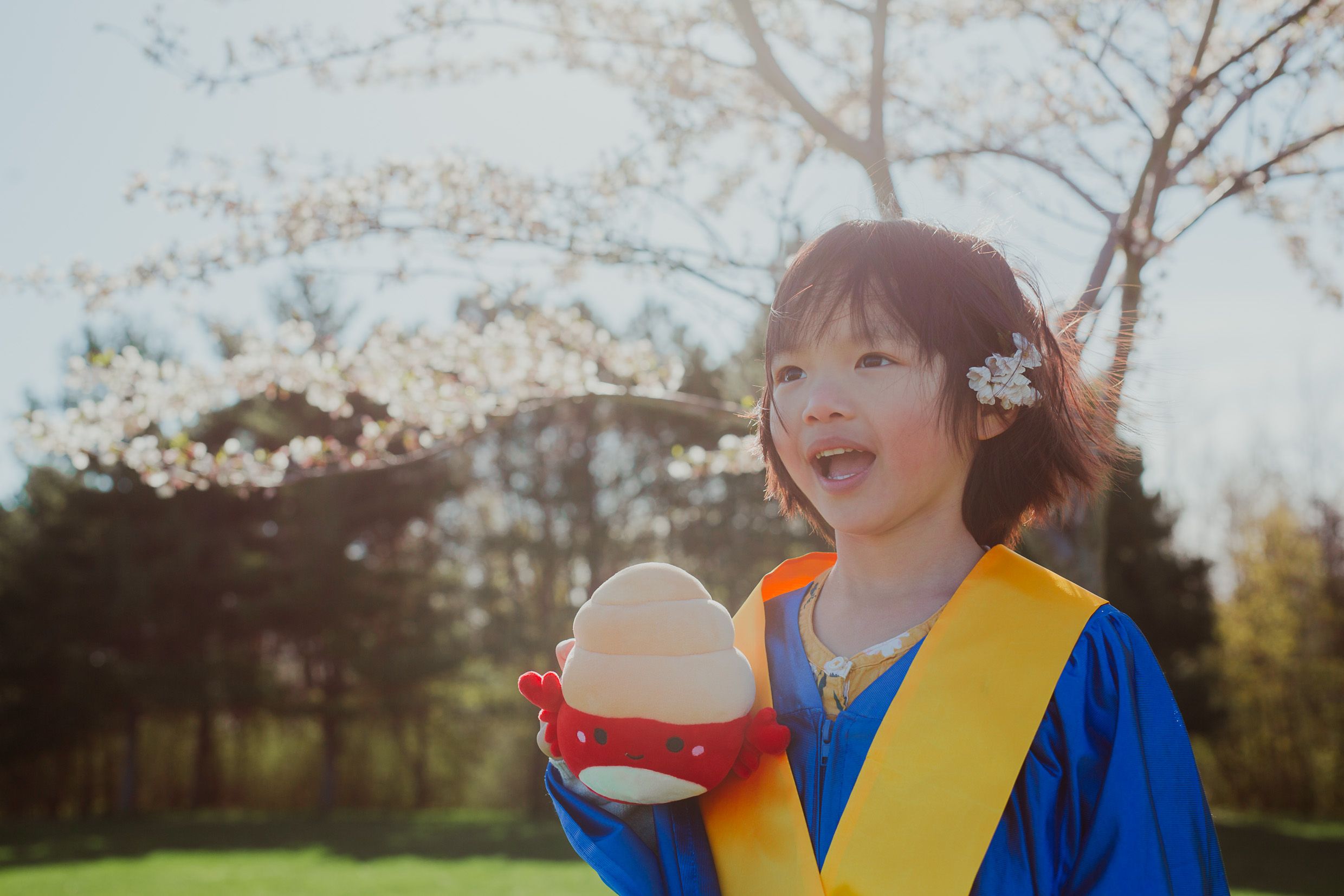A person in blue and yellow graduation attire holds up a round ball or diploma case under blooming spring trees.