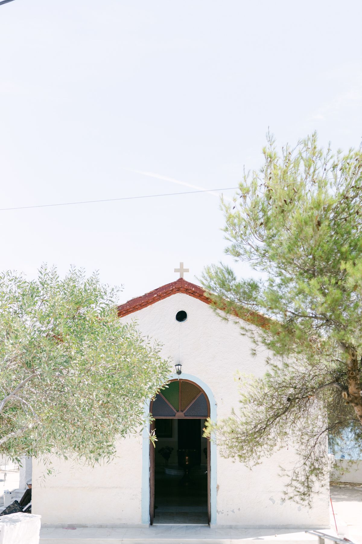 Small white Mediterranean chapel with arched blue doorway and wooden cross detail framed by olive trees against pale sky.