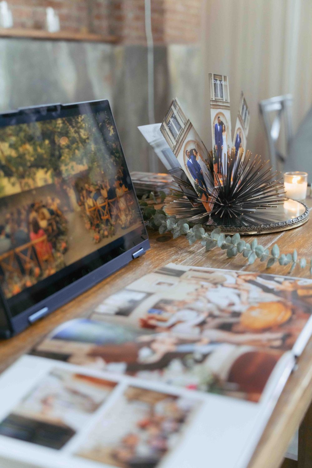 Event photo display with laptop and decorative fan-style photo arrangement on wooden table. By FORGET ME NOT ARTS