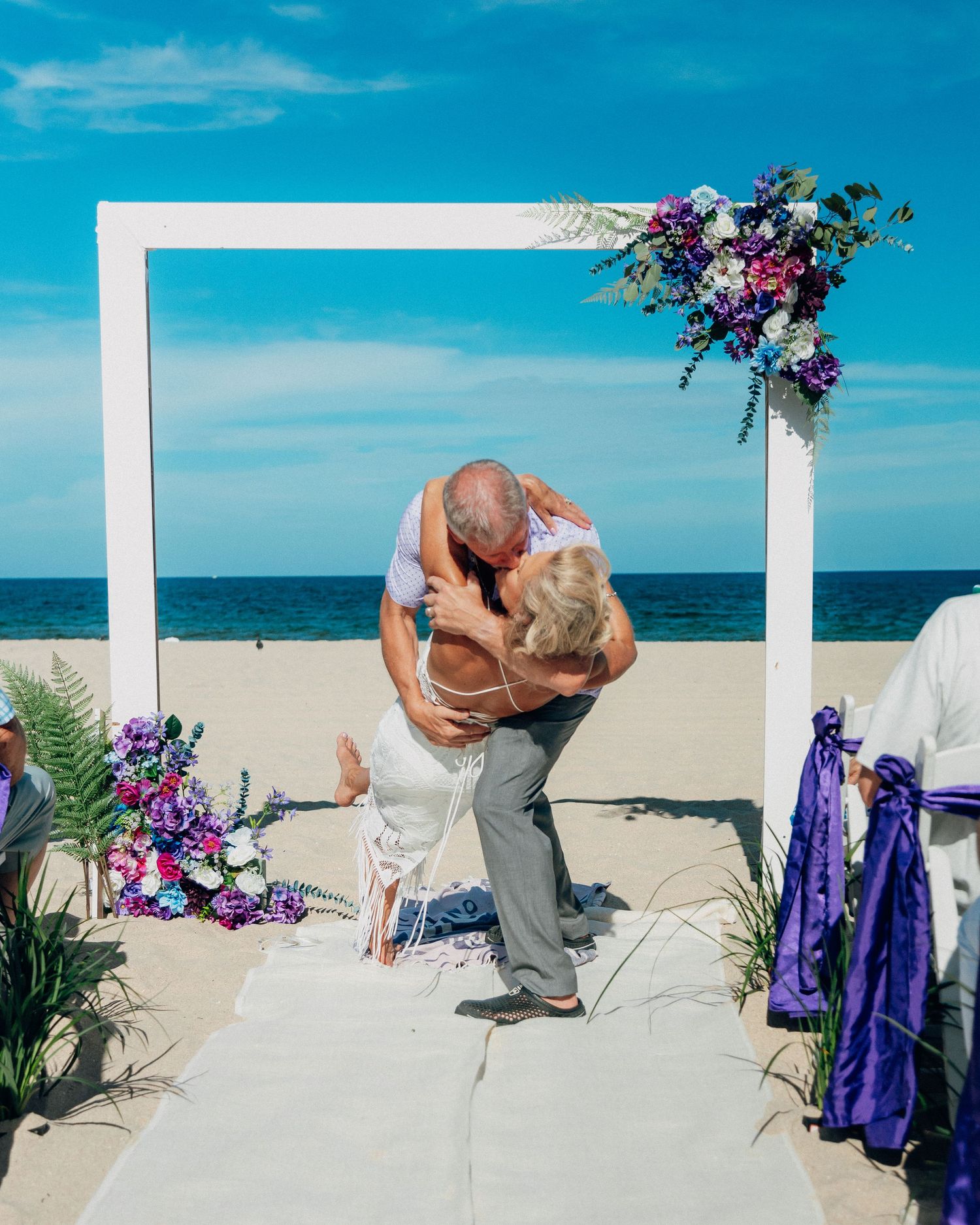 A romantic beach wedding ceremony features a white arch decorated with purple flowers against the ocean.