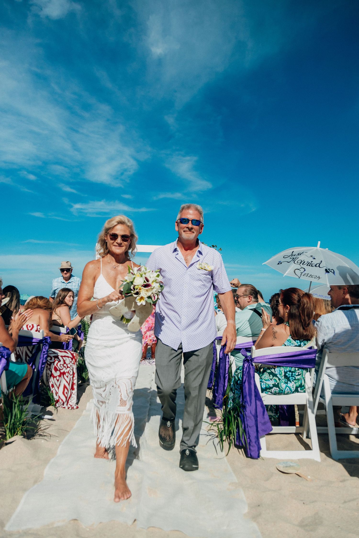 A beach wedding celebration with couple walking down aisle decorated in purple and white decor.
