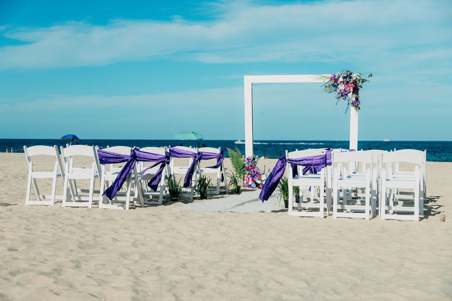 Beach wedding setup with white chairs adorned with purple sashes and simple white arch on sandy shore.
