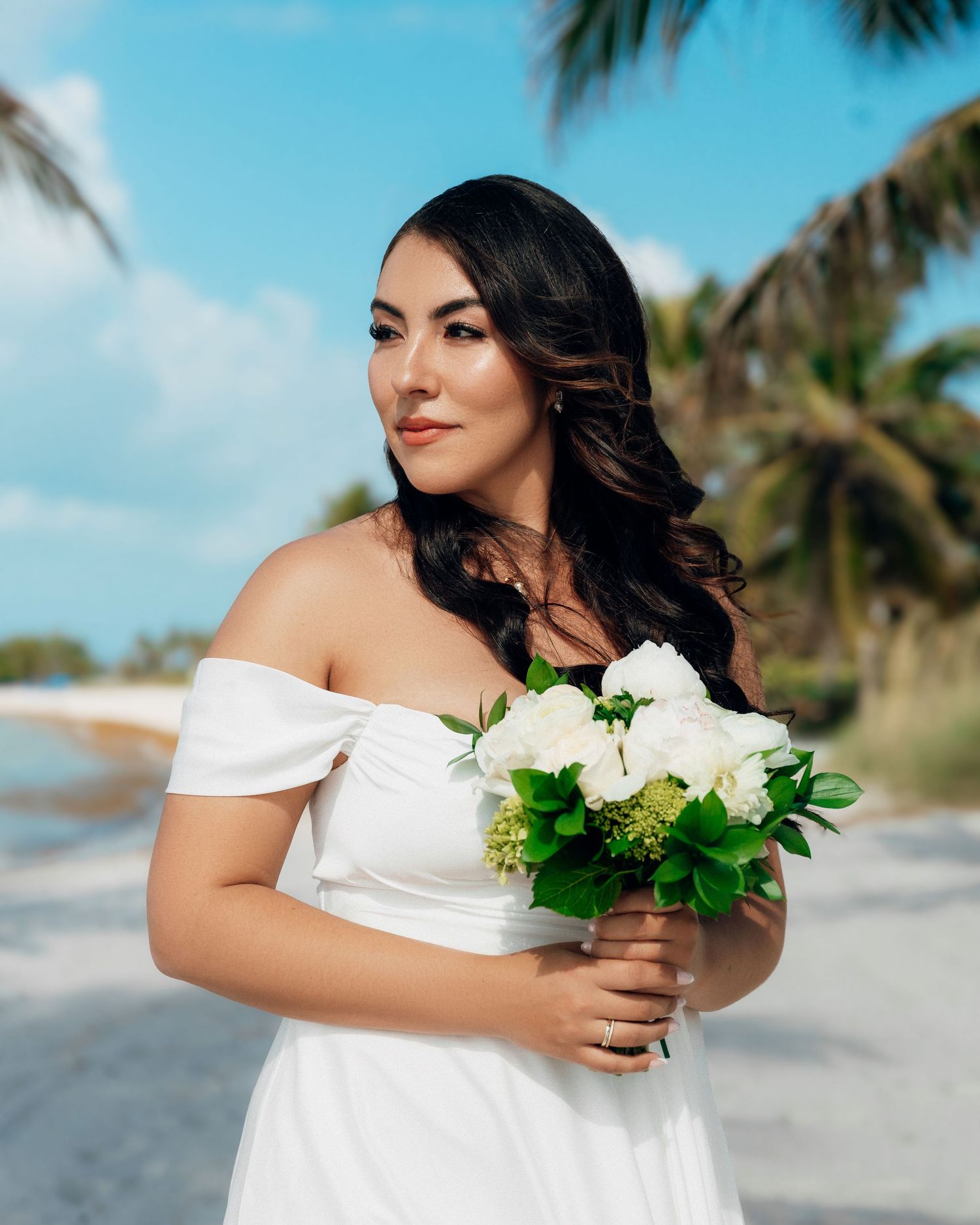 A person in an off-shoulder white dress holds a white rose bouquet on a tropical beach with palm trees and blue sky.
