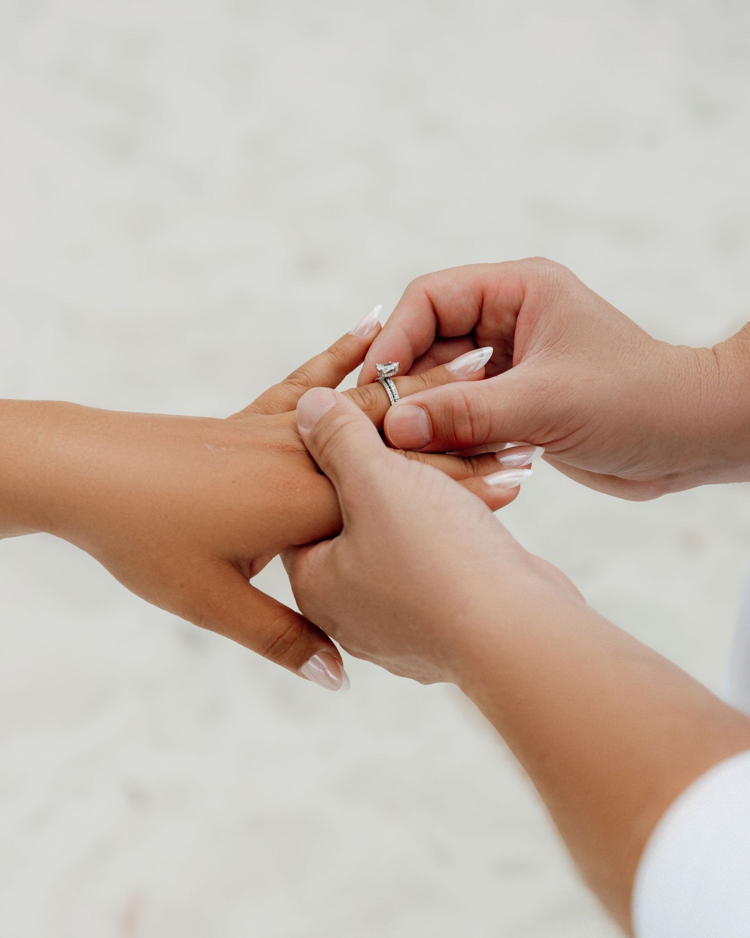 A close-up shot of hands placing an engagement ring on a finger against a light background.