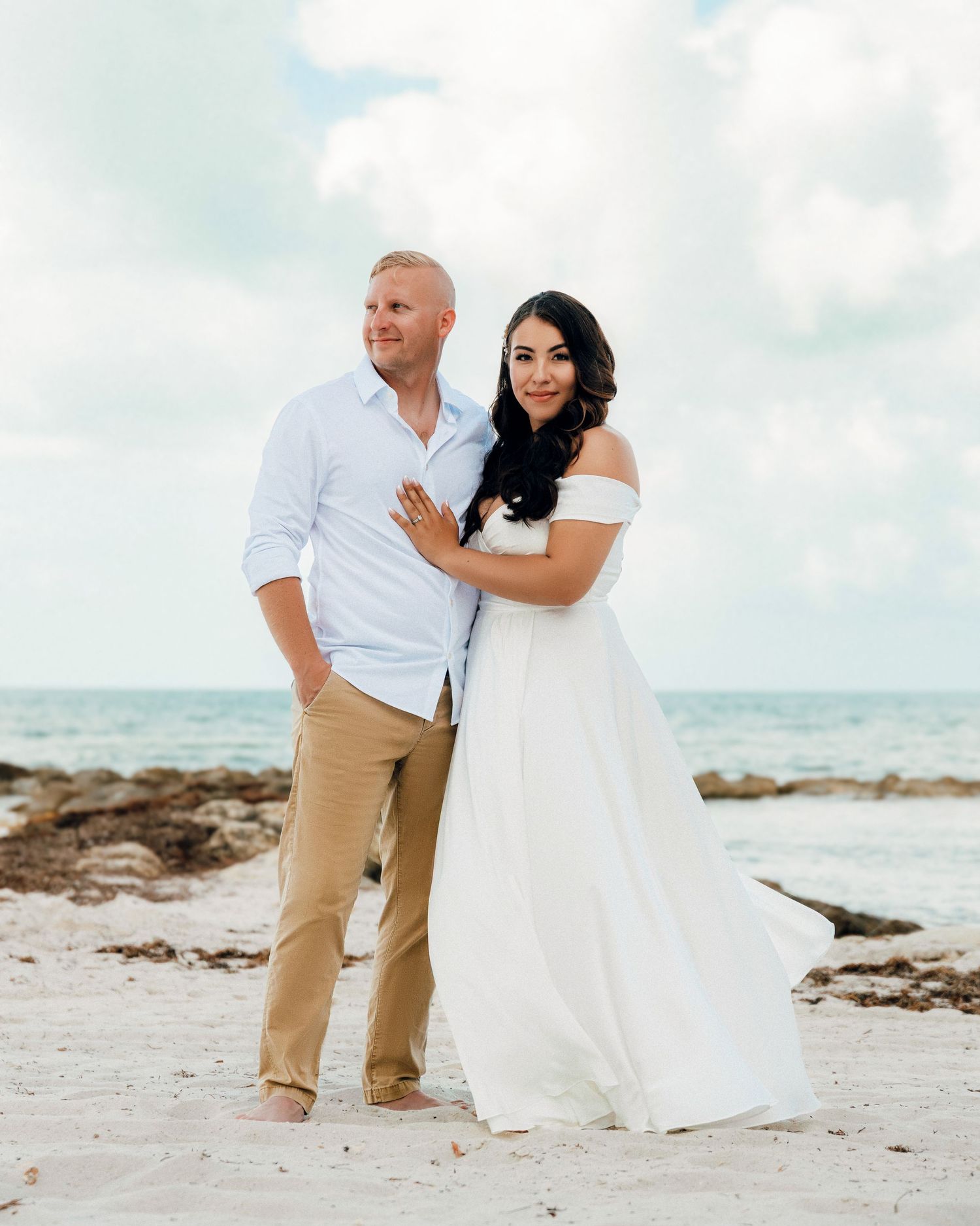 A couple poses together on a tropical beach with white sand and ocean waves in the background.