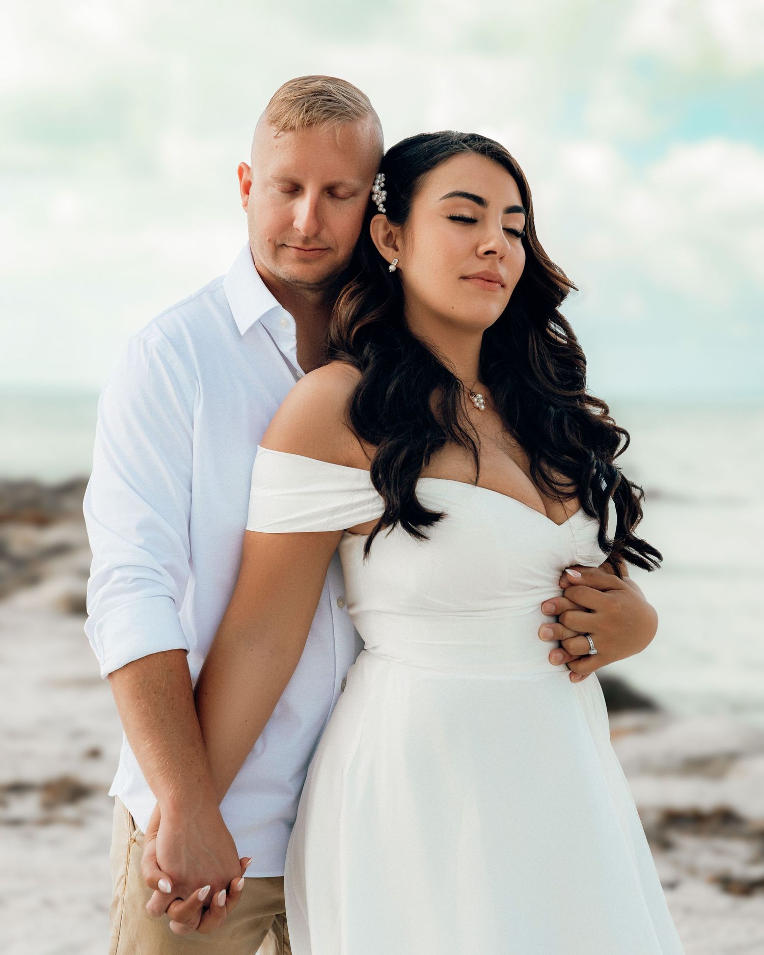 A romantic couple embraces on a beach, with one person wearing a white off-shoulder dress and the other in a light button-down shirt.