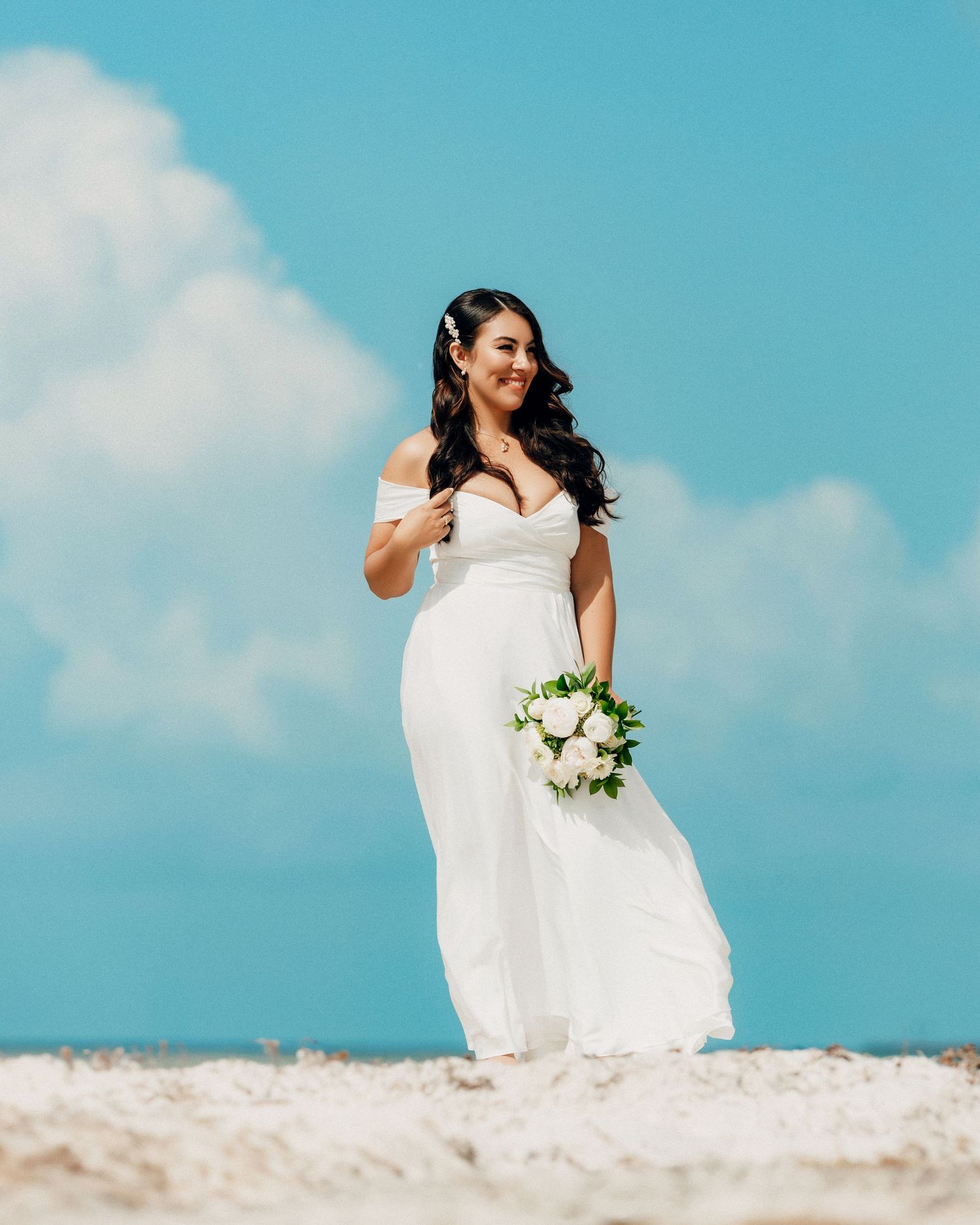 A bride in a flowing white off-shoulder dress holds a bouquet while standing on a sandy beach against blue skies.