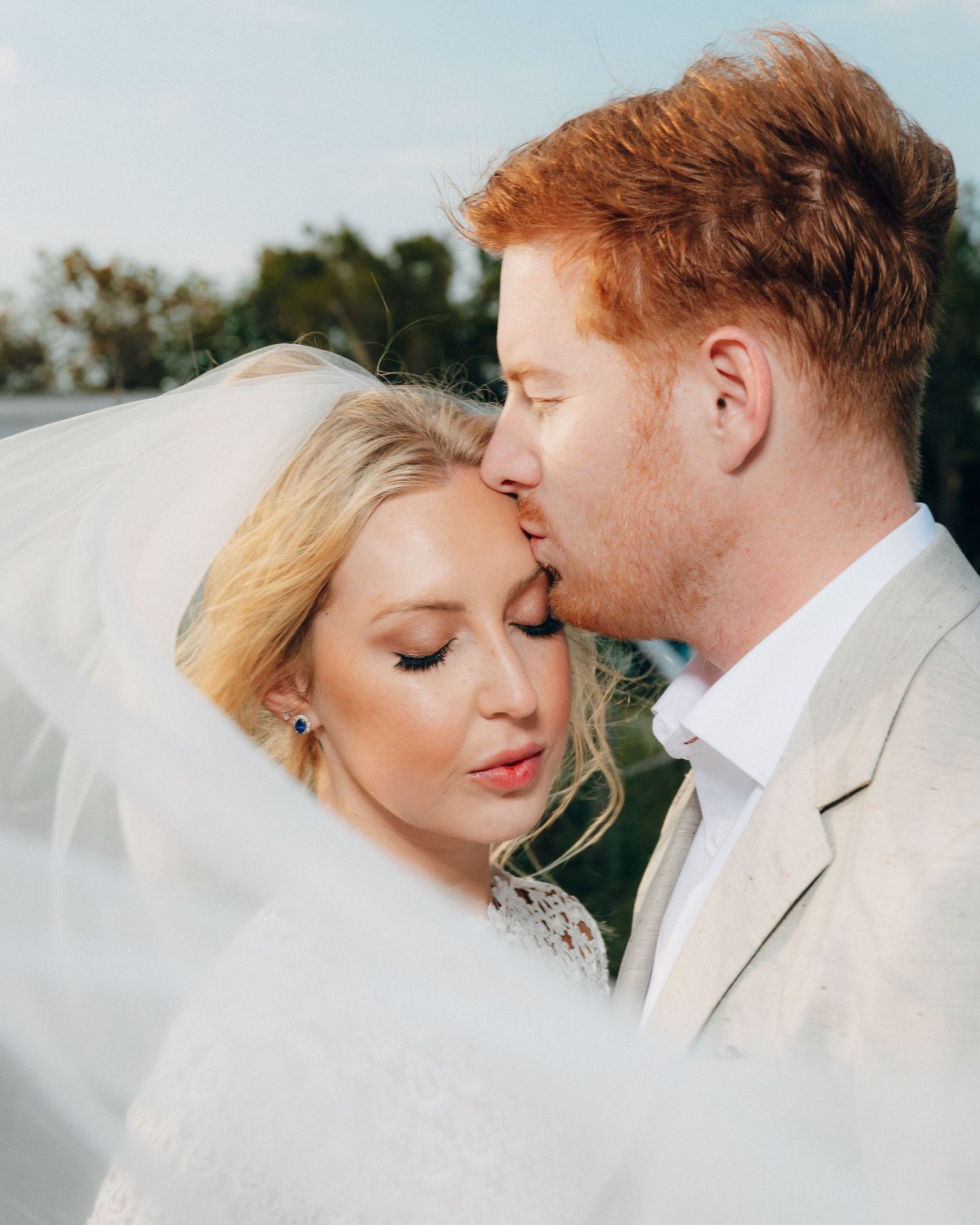 A romantic wedding photo of a couple sharing a tender moment under a veil outdoors.