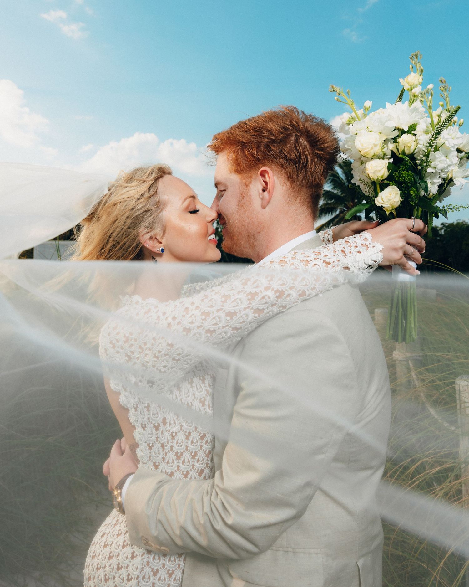 A romantic wedding kiss between newlyweds outdoors with a flowing veil and white floral bouquet.