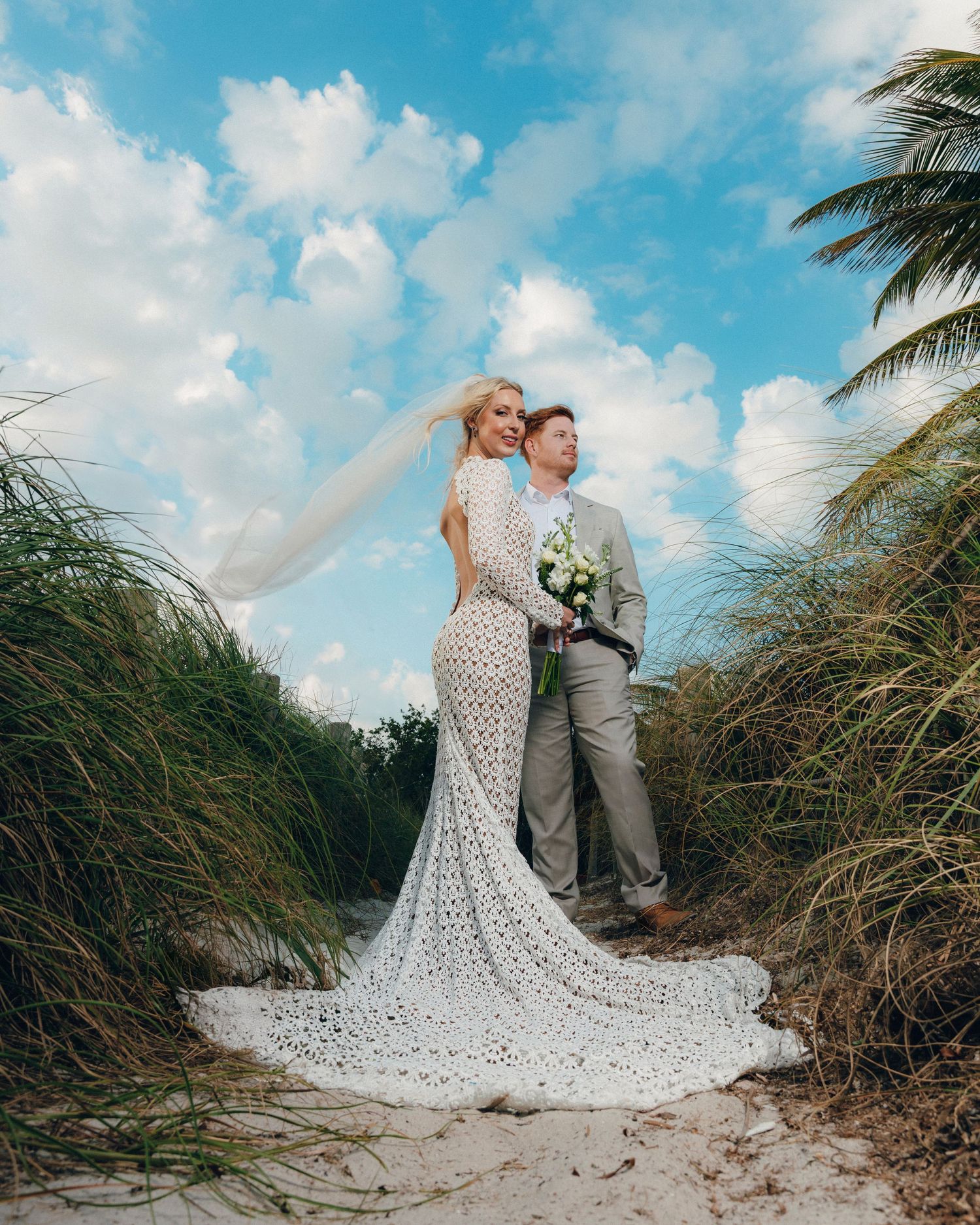 A couple poses on a sandy beach path between tall grass and palm trees on their tropical wedding day.