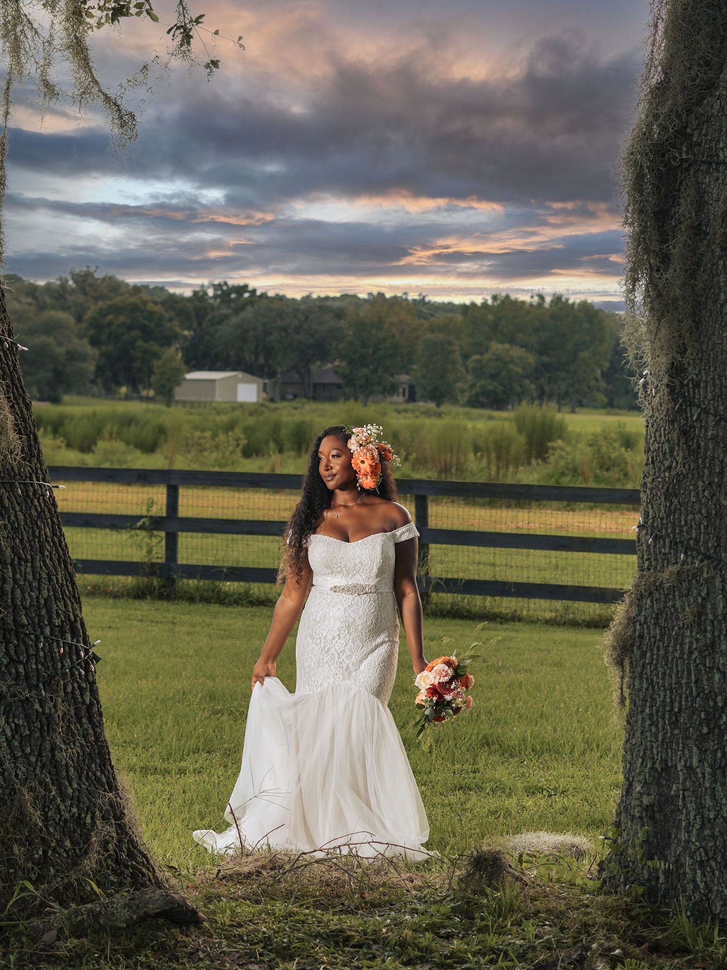 A bride in a white mermaid dress poses between trees at sunset with a rustic fence backdrop.