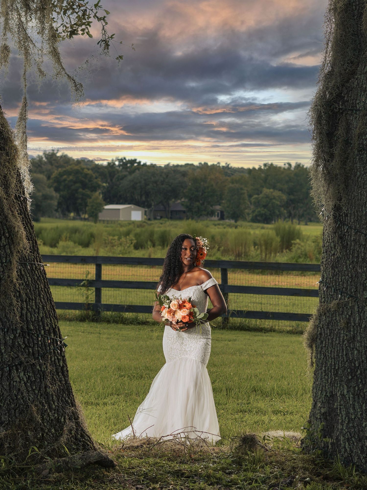 A bride holds a coral bouquet while standing between mossy trees at sunset with a fence in the background.