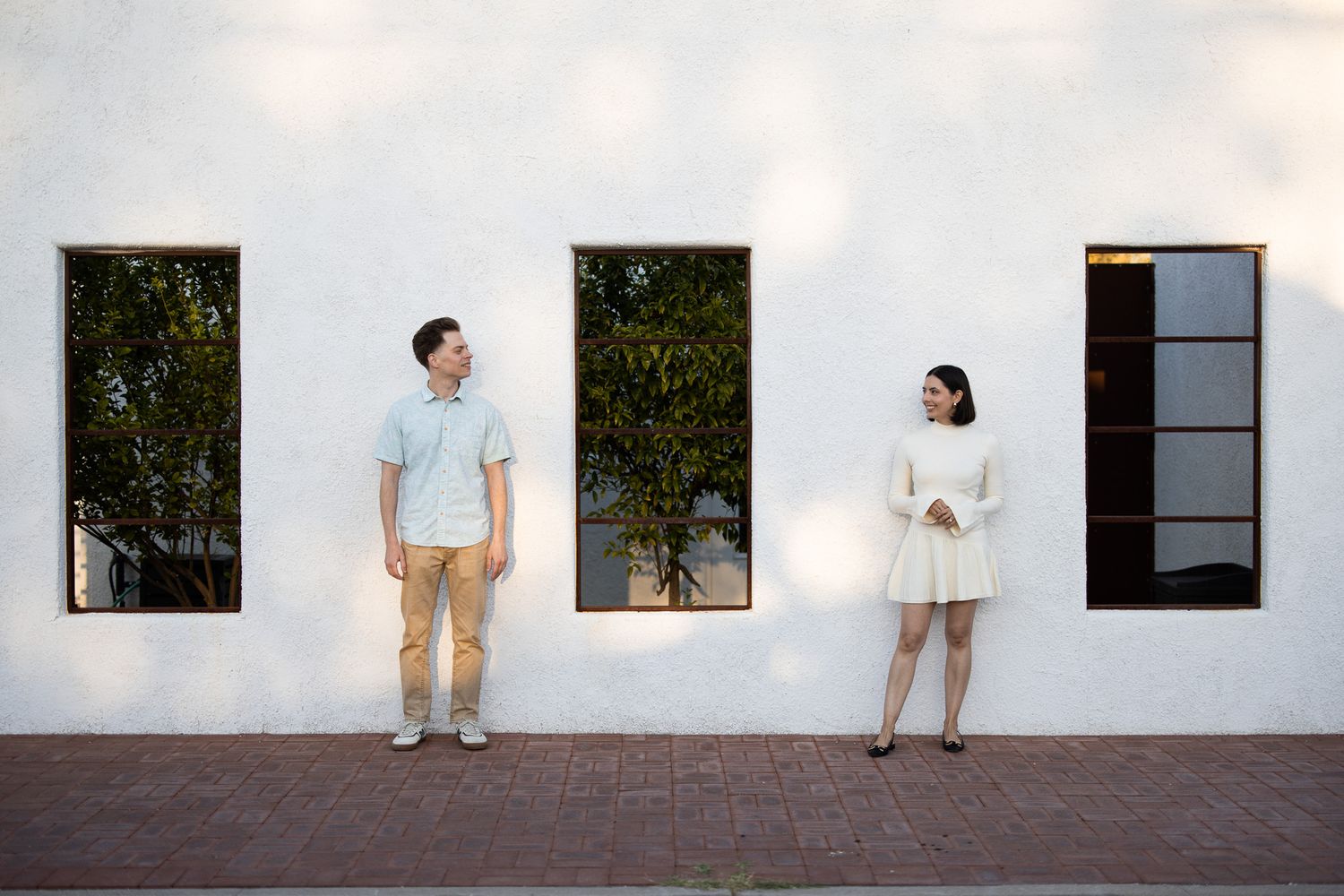 Two people stand distanced apart against a white wall with dark window frames and greenery, creating a minimalist scene.