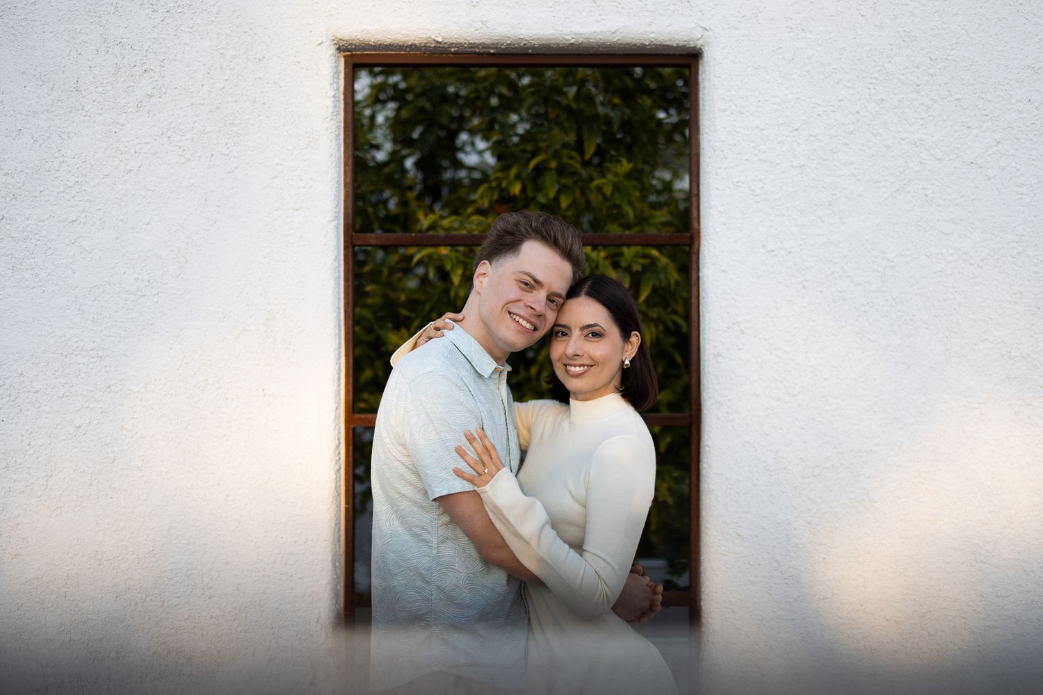 A romantic outdoor portrait of a couple embracing in white sweaters against a leafy green background.