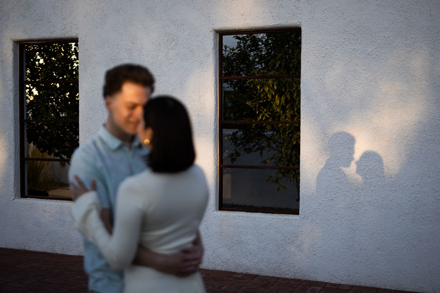 Silhouetted couple embracing creates romantic shadows on white wall between two dark windows at sunset.